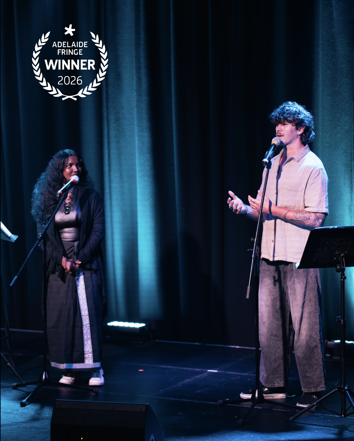 Two performers on stage at the Adelaide Fringe 2026, with one man speaking into a microphone and a woman listening, set against dark curtains and stage lighting.