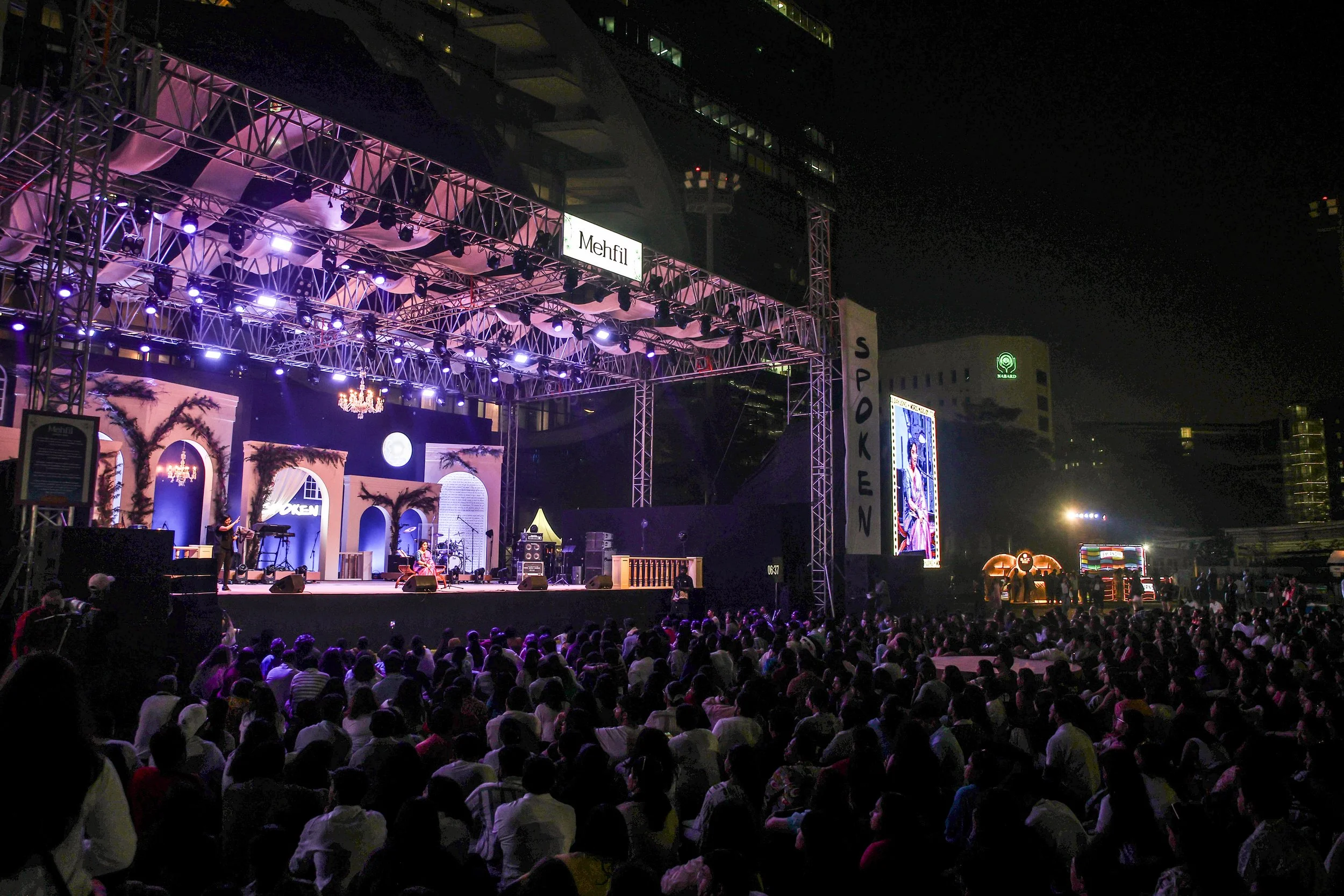 A nighttime outdoor concert stage with purple lighting, decorated with arches and palm leaves, with a crowd of seated people watching a performance. There are large screens displaying performers, and an illuminated sign says 'Mehfil'.
