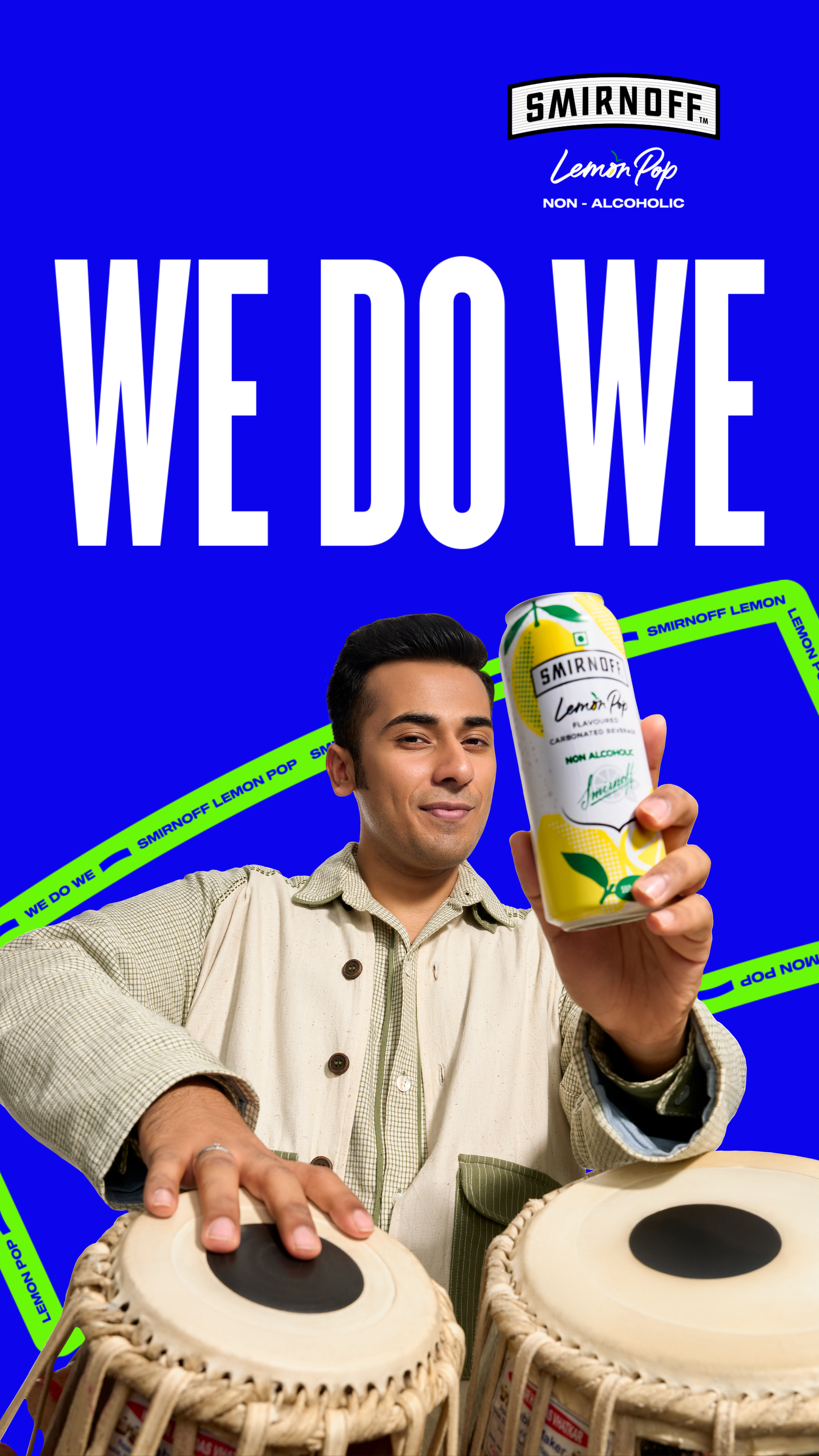 A man wearing a jacket and shirt is holding a can of Smirnoff Lemon Pop non-alcoholic beverage. He is smiling. The background features large white text on a blue background that says "WE DOWE". There are green tape strips with blue text that say "WE DOWE SMIRNOFF LEMON POP".