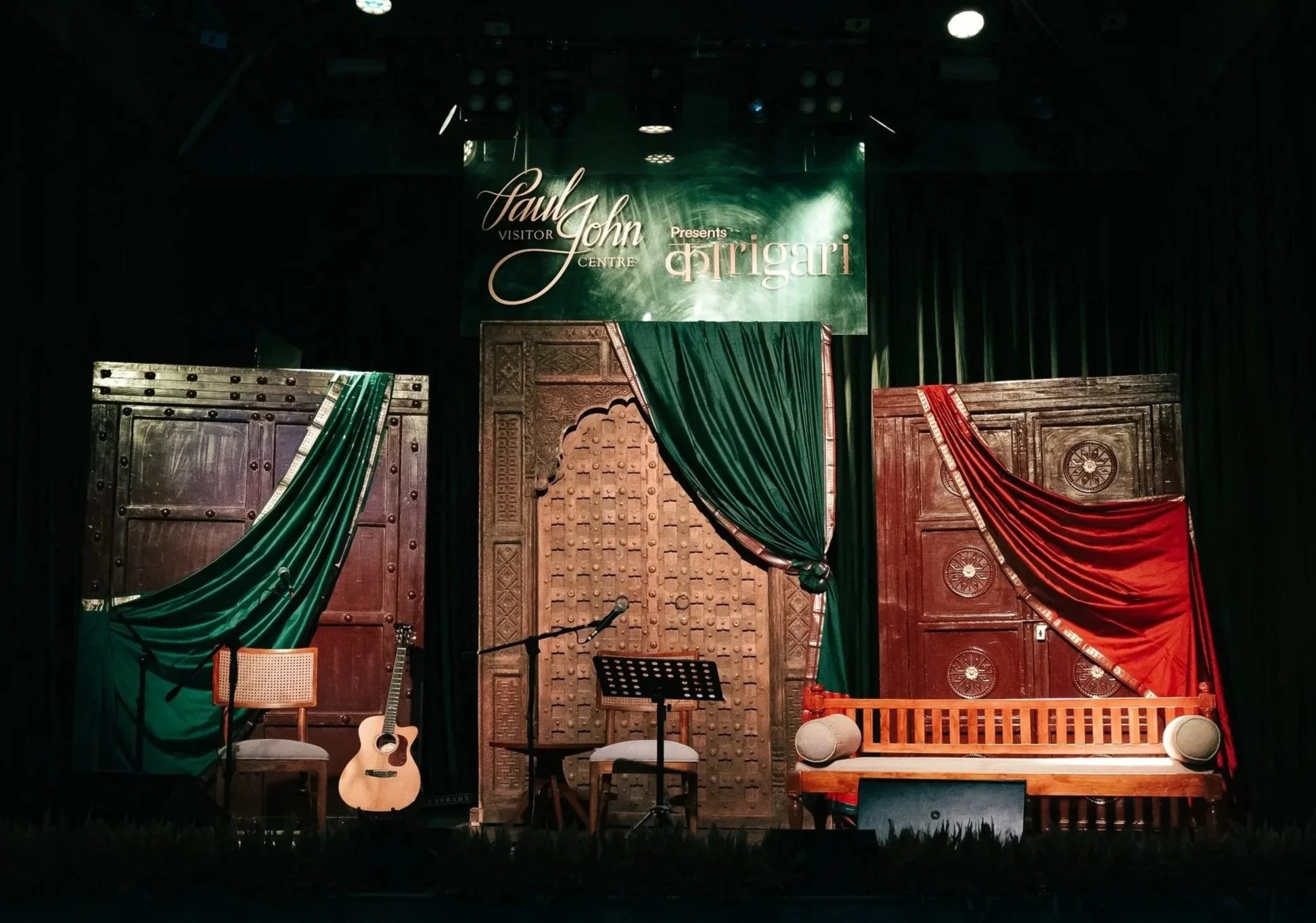 Stage setup with musical instruments and seating, featuring wooden doors and green and red draped curtains, in a cultural performance venue.