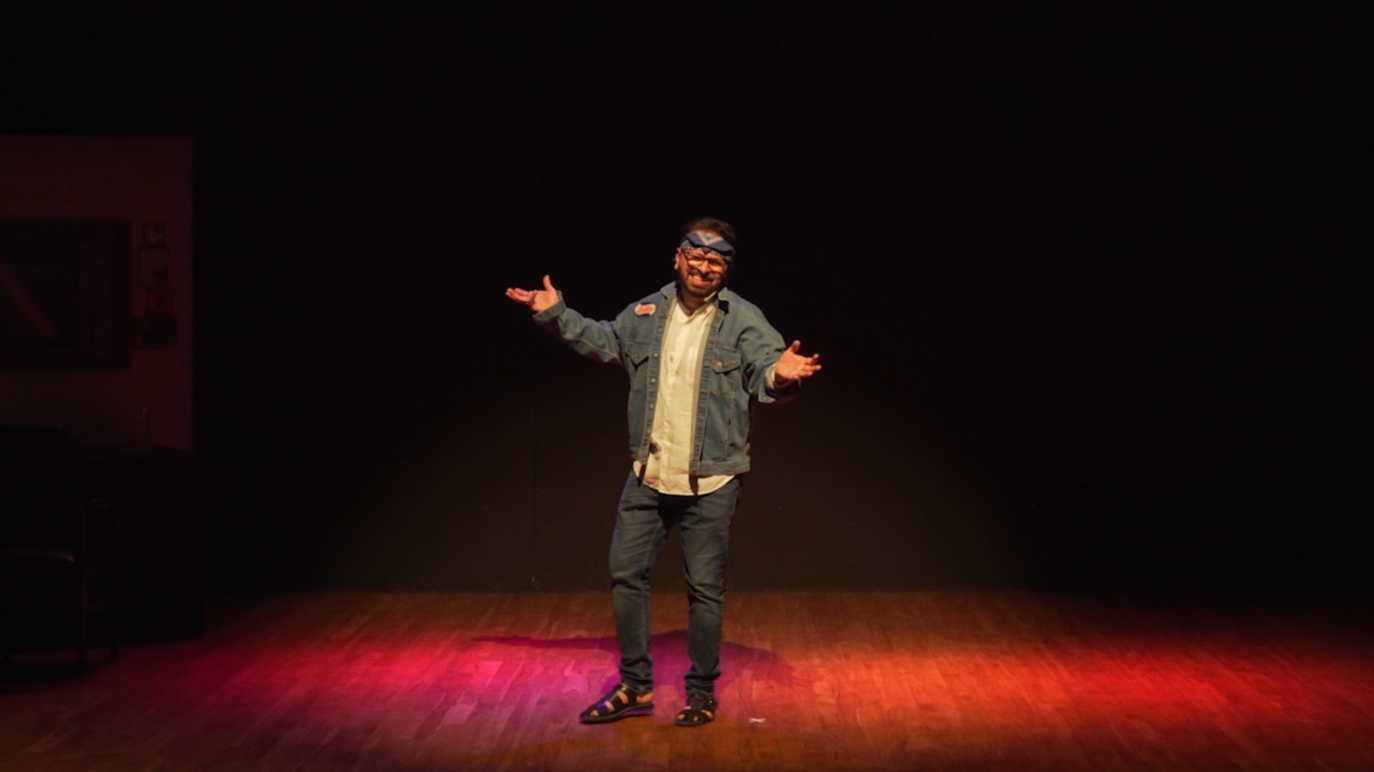 A man standing on a stage with a black background, wearing a denim jacket, a party hat, and sandals, smiling with arms outstretched.