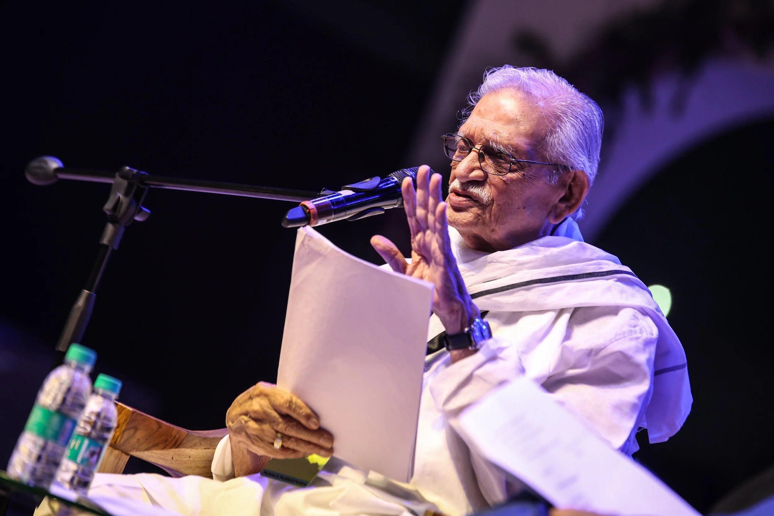 An elderly man with white hair and glasses, dressed in traditional Indian attire, is speaking into a microphone at a stage with a black background, with water bottles on the table in front of him.