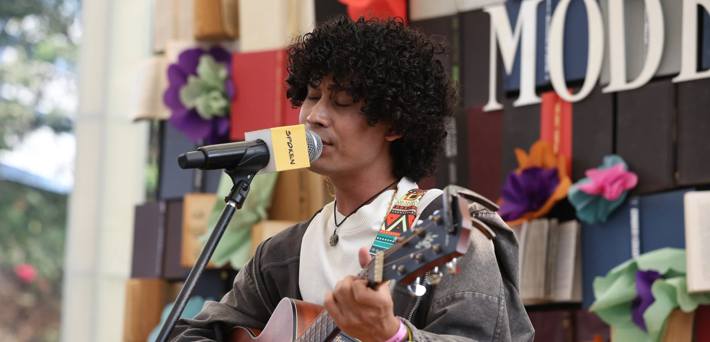 A young person (Maan Panu) with curly hair singing into a microphone labeled 'SPROKEN' while playing an acoustic guitar, in a room decorated with colorful paper flowers and books.