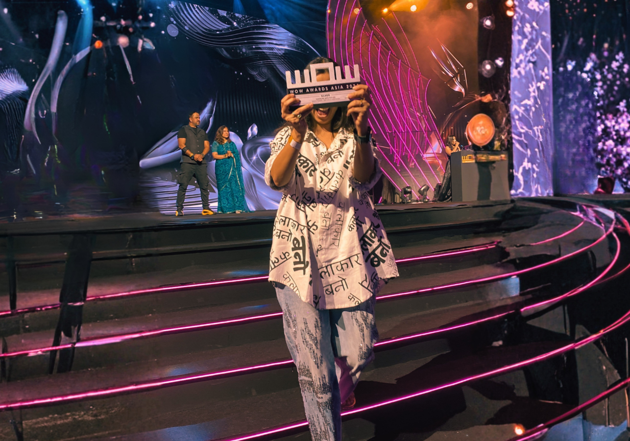 A woman on stage holding up a WOW Awards trophy, smiling. She is wearing a printed outfit with black and white text. In the background, there are three people, two men and one woman, with colorful abstract design on the stage backdrop, and a DJ station to the right. The stage has curved stairs with pink lighting.