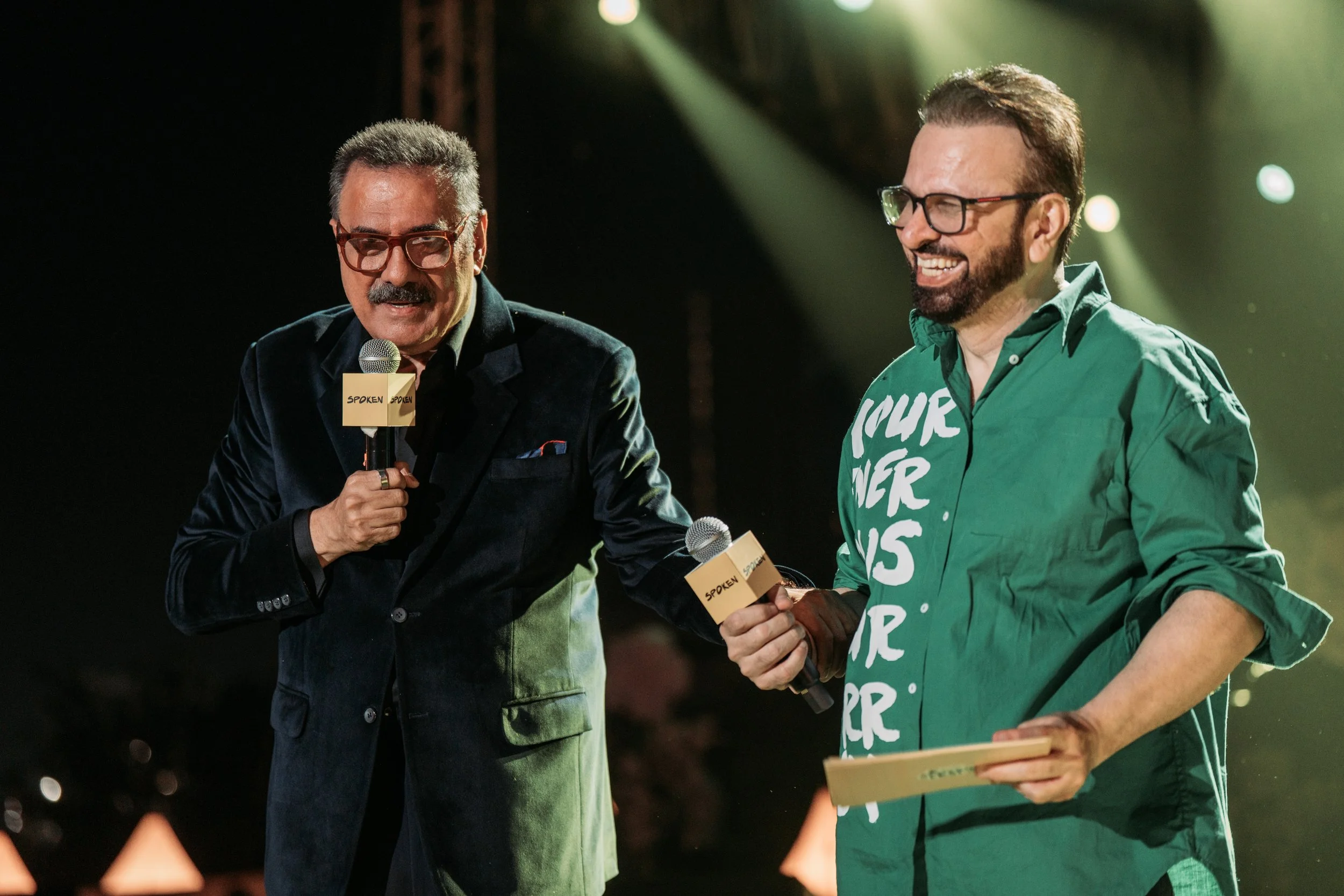 Boman Irani & Roshan Abbas on stage, smiling and holding microphones with 'SPOKEN' labels, engaging in conversation during an event.