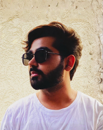 A man with dark hair and a beard wearing sunglasses and a white T-shirt, standing against a textured beige wall.
