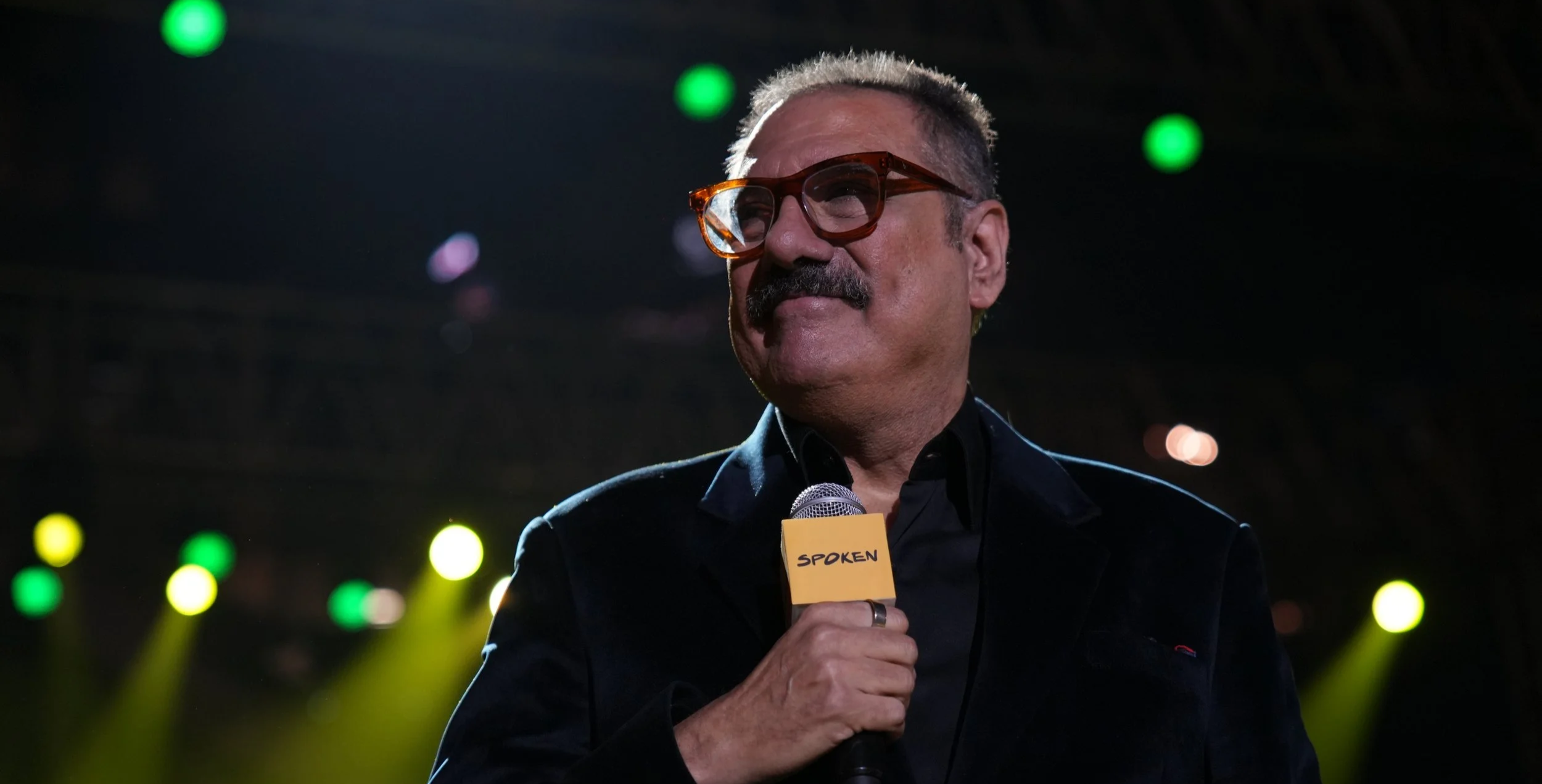 A man (Boman Irani) with gray hair and a mustache wearing glasses and a black shirt, holding a microphone labeled 'Spoken,' standing on a stage with colorful lights in the background.