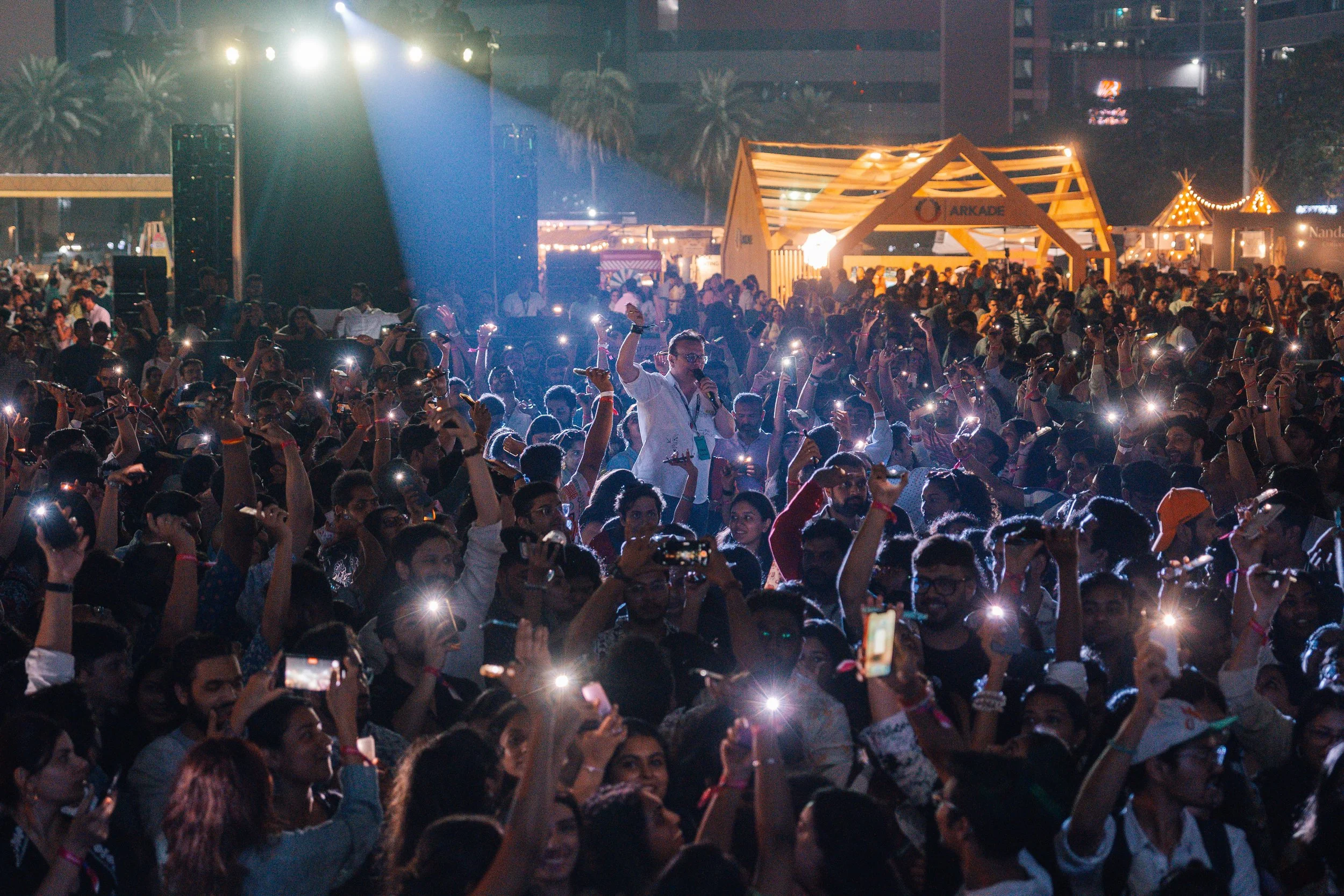 A large crowd of people gathered at night, many holding up their phones with flashlight on, in front of a stage with bright lights and a man speaking into a microphone. There are booths and structures in the background, including a brightly lit wooden structure labeled 'Arkade' and decorative tents.