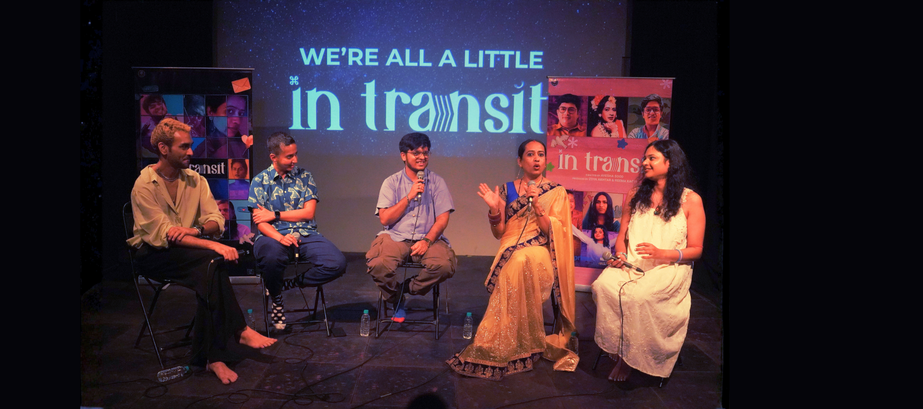 Five people sitting on a stage participating in a panel discussion with a large screen behind them displaying the words "WE'RE ALL A LITTLE in transit." Two women and three men are engaged in conversation, holding microphones, with posters and banners related to the event visible in the background.