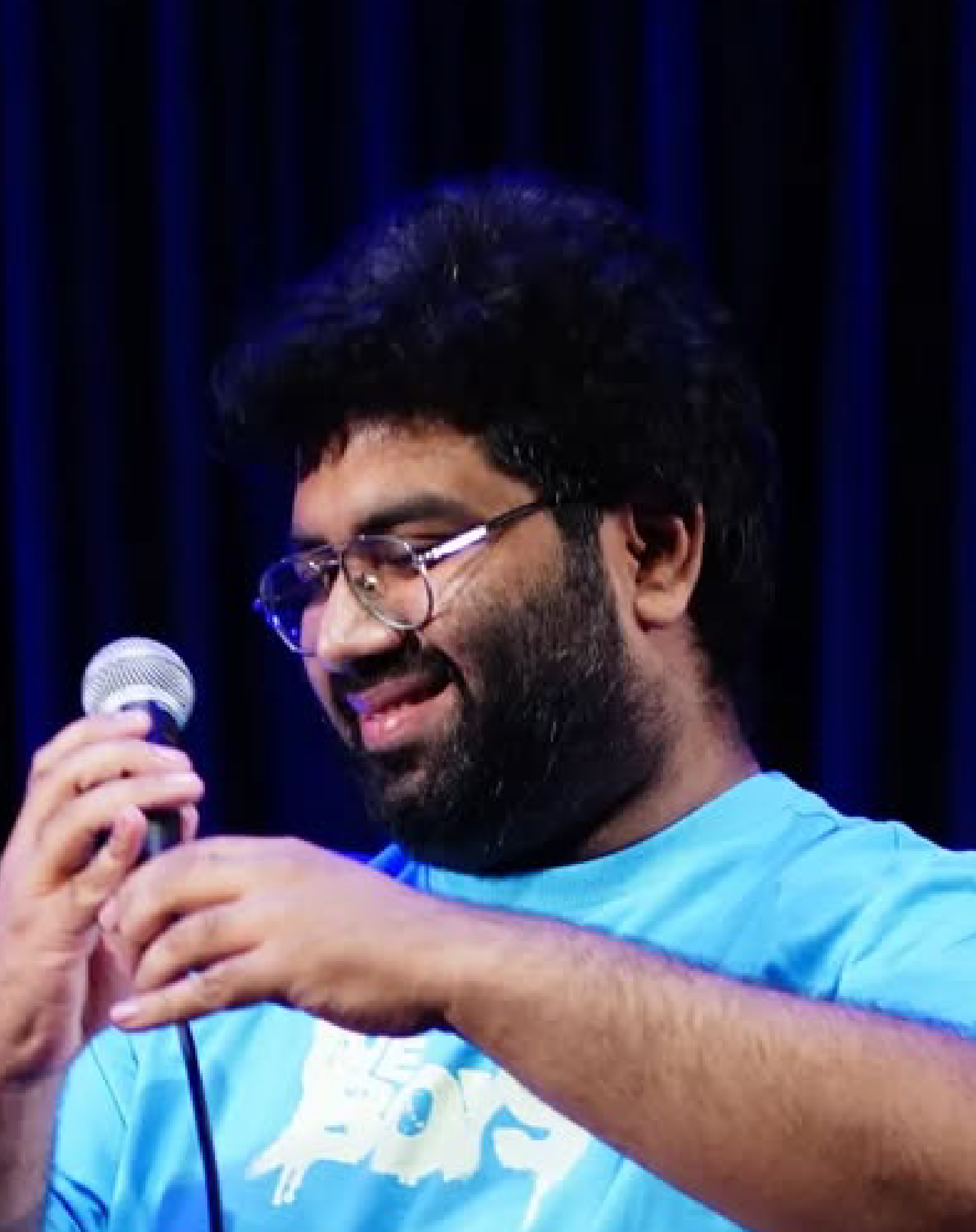 A young man with glasses and a beard holding a microphone on stage, smiling, with dark curtain backdrop.