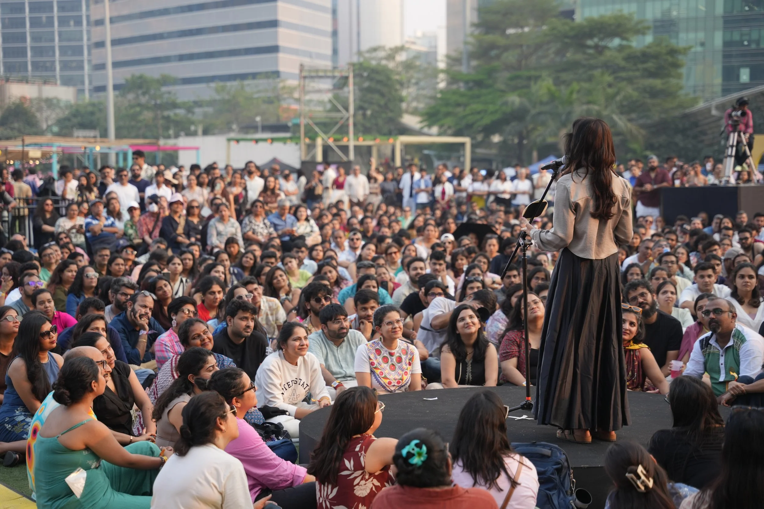 A woman (Sriti Jha) with long brown hair standing on a small stage speaking into a microphone in front of a large seated crowd outdoors in an urban area.