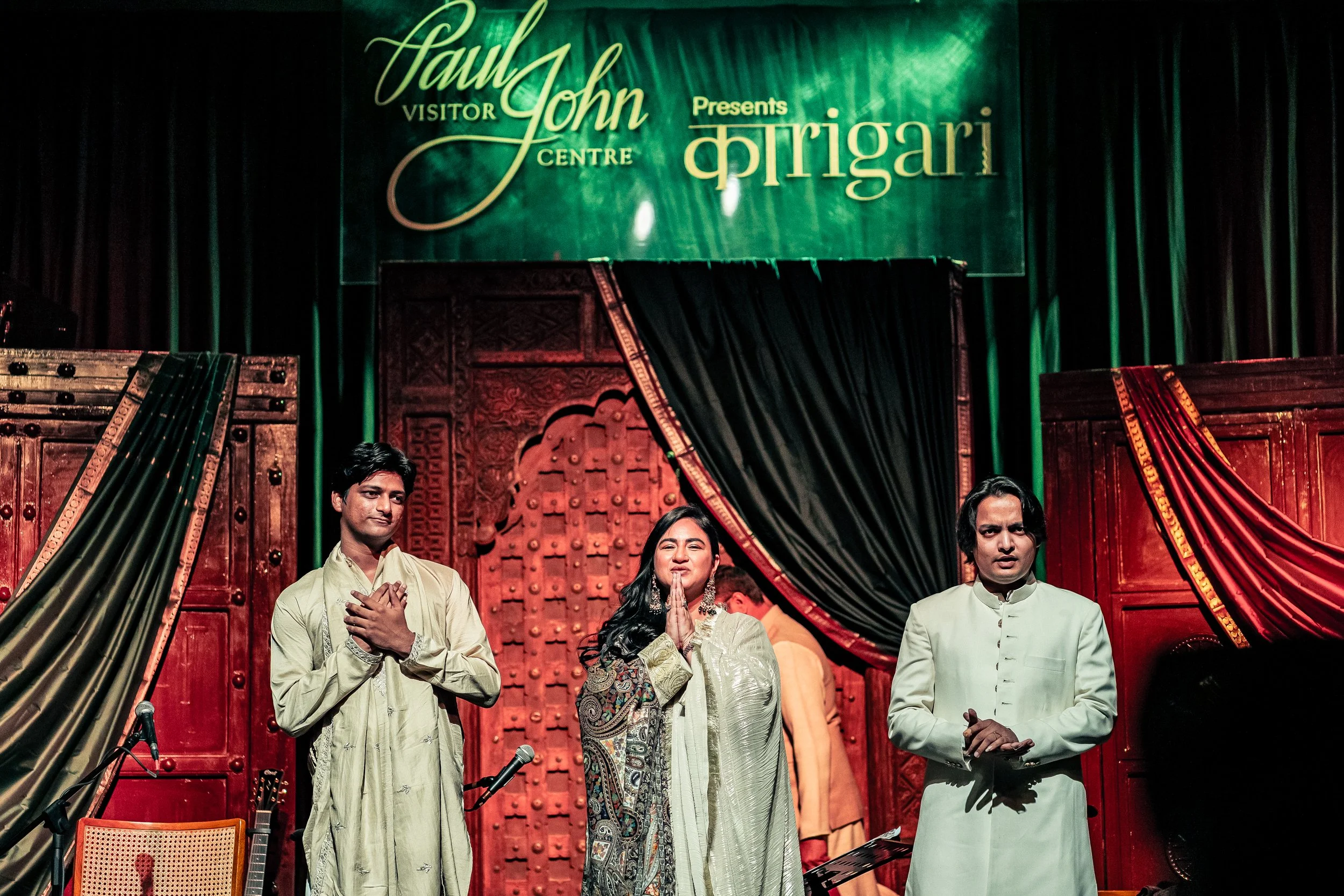 People standing on stage during a performance, with a large green banner behind them that reads 'Paul John Visitor Centre Presents Kaarigari'.