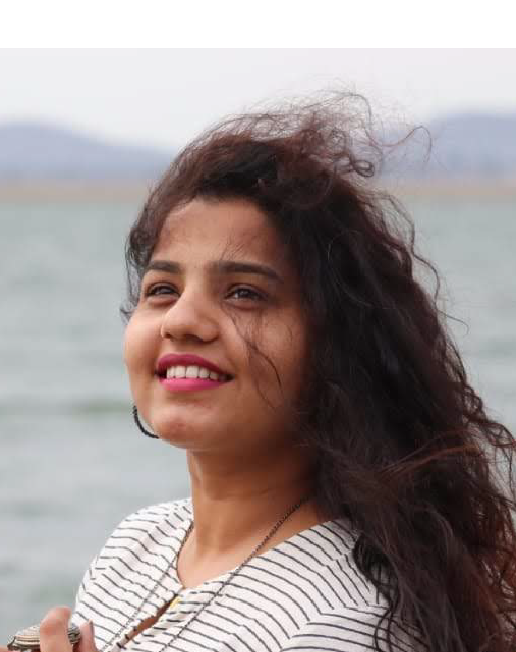 A woman smiling with wind-blown hair standing by the water with a distant shoreline and mountains in the background.