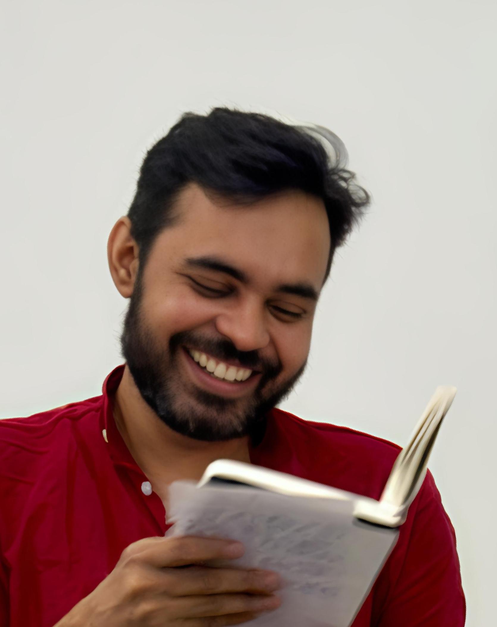 A man smiling and looking down at a notebook he is holding in his right hand, wearing a red shirt against a plain white background.