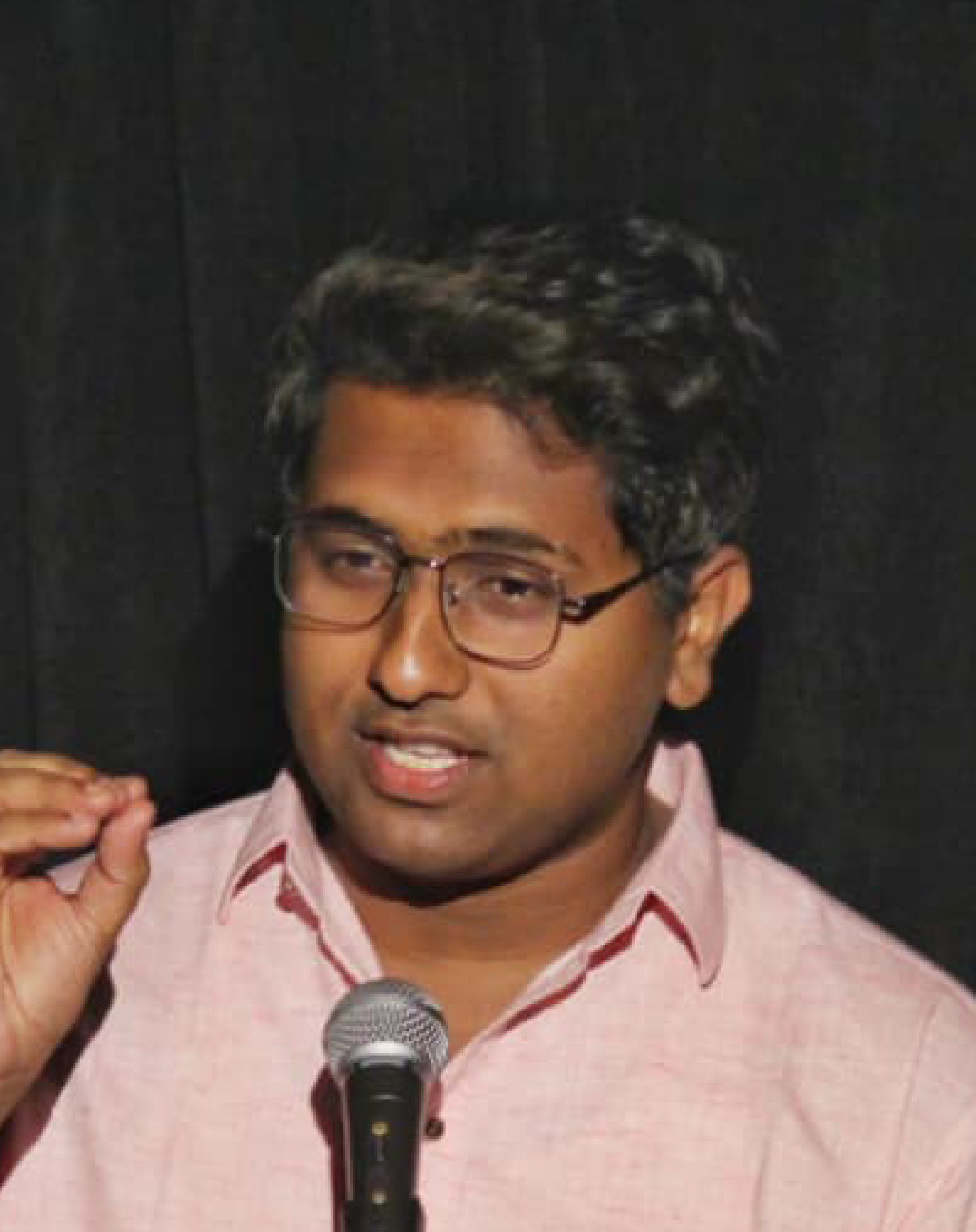 A man with glasses, wearing a pink shirt, speaking into a microphone and gesturing with his hand.