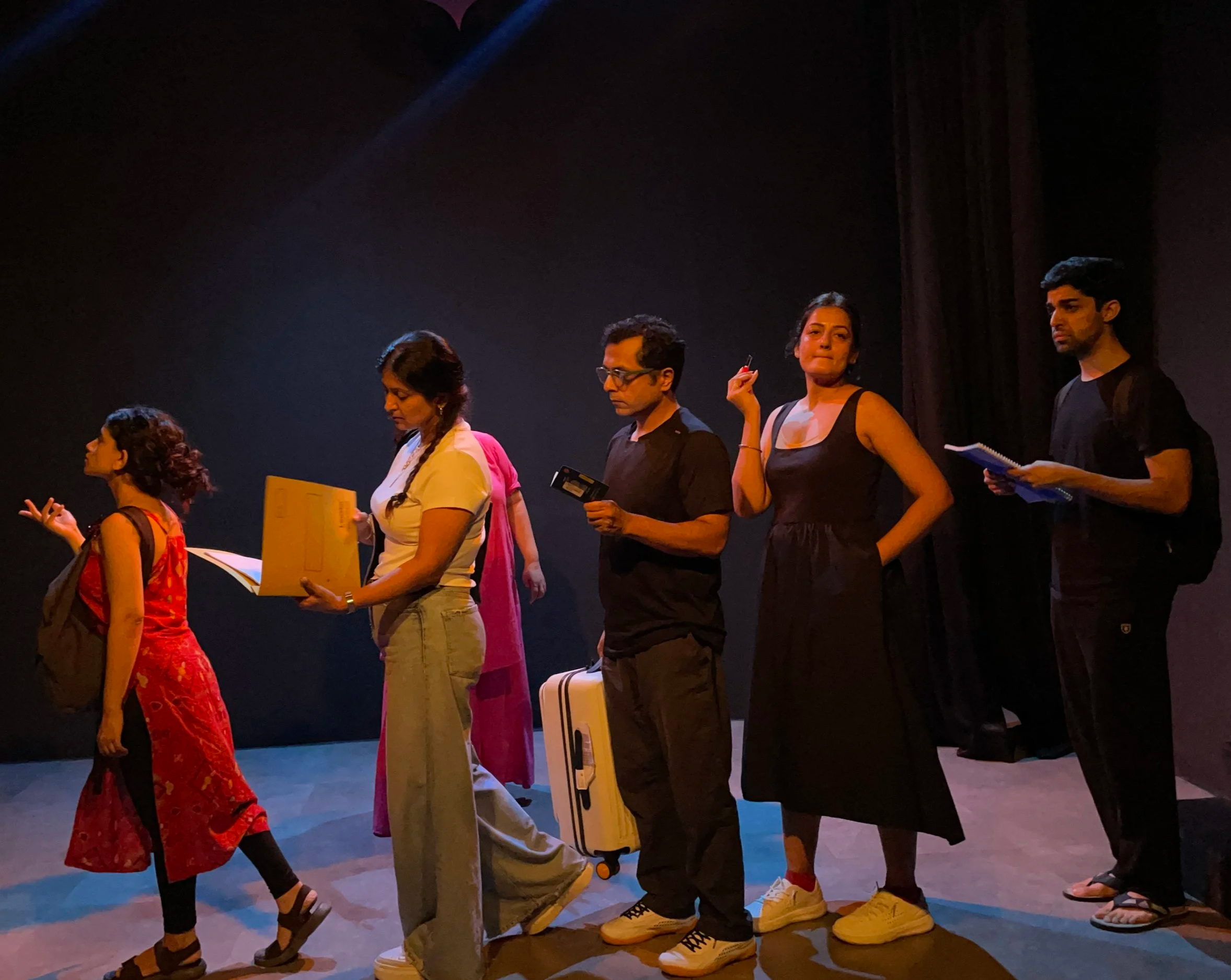 A diverse group of six actors or performers standing in a line on stage, engaged in a theatrical performance. The stage is dimly lit with a dark background.