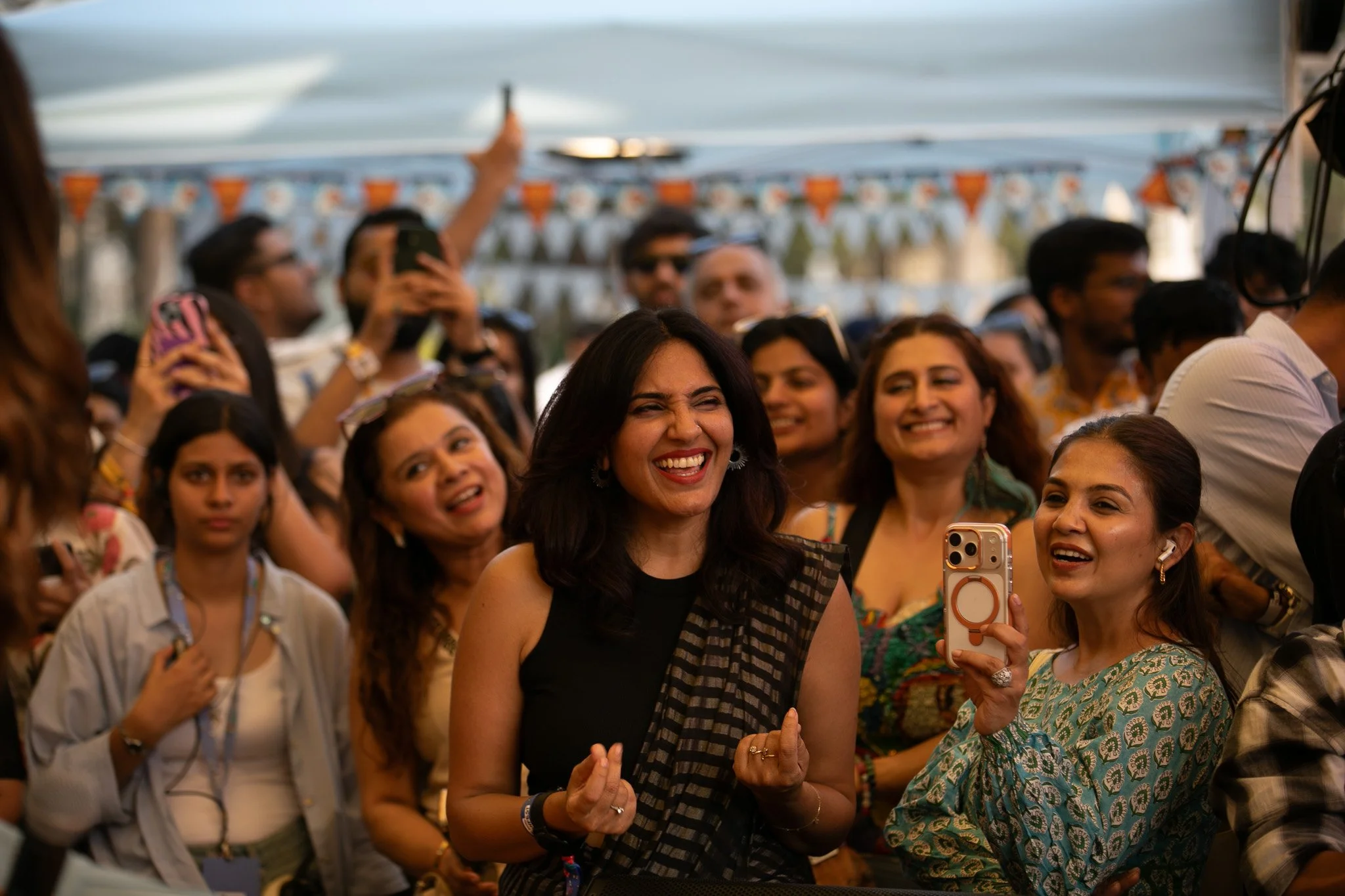 Group of women at an outdoor event, smiling and taking photos, with a woman in black and black striped saree in the center.