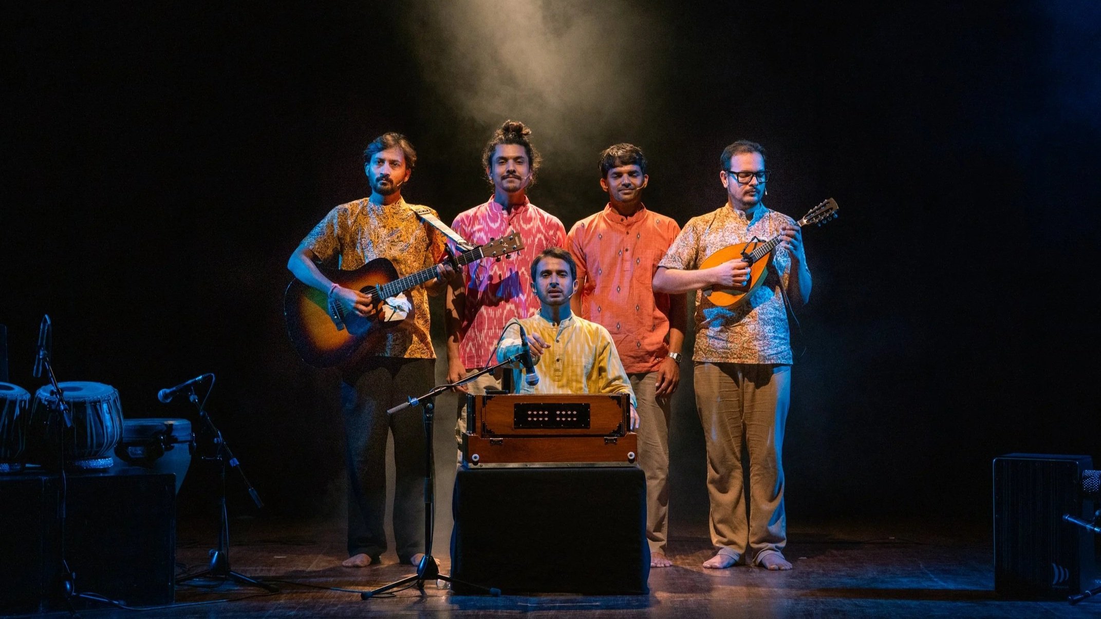 Six men are performing on stage outdoors, with three standing and two sitting. The men are playing guitars and sitar, and the stage is dimly lit with a dark background.