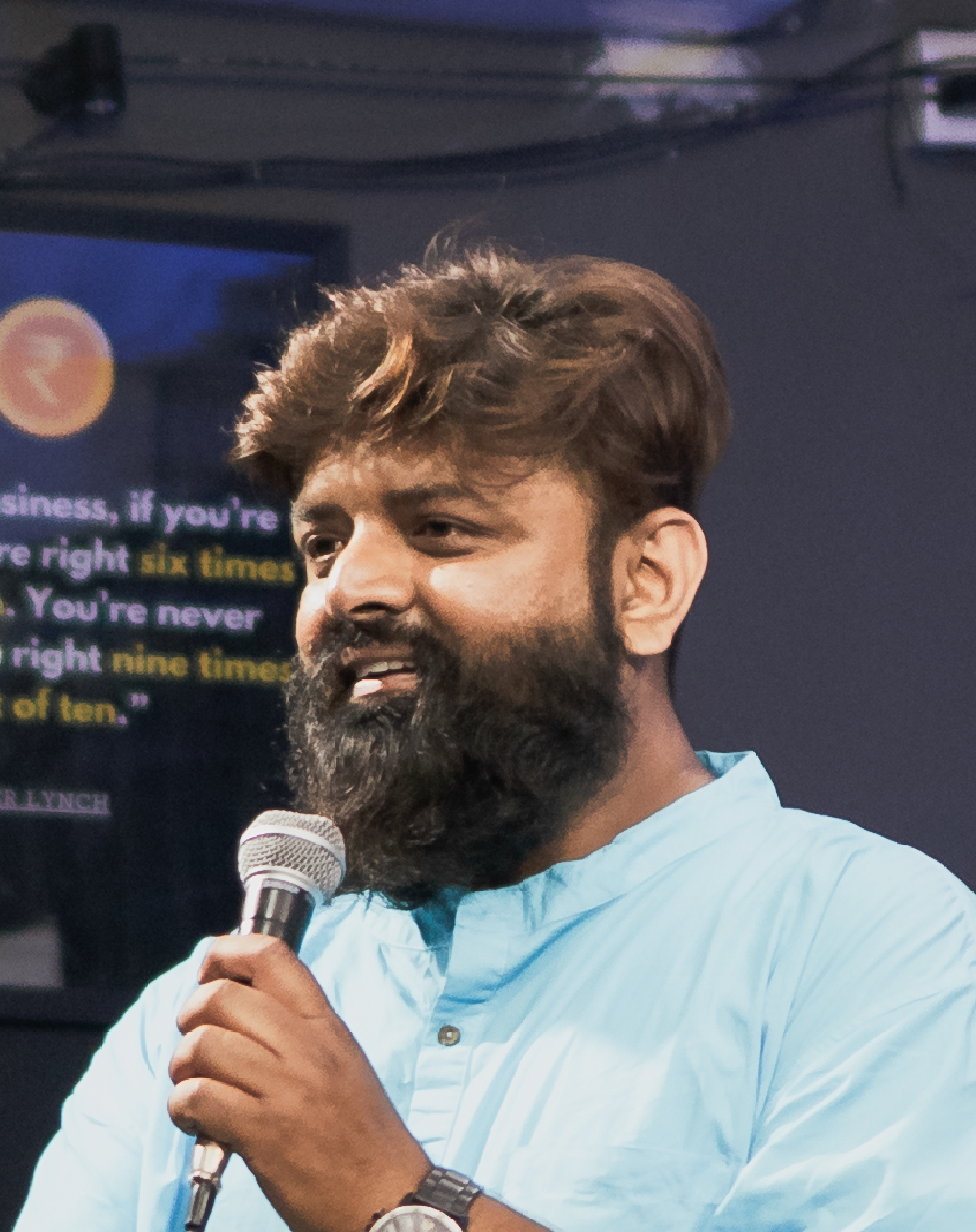 Man with a beard and mustache, wearing a light blue shirt, speaking into a microphone during a presentation or speech.