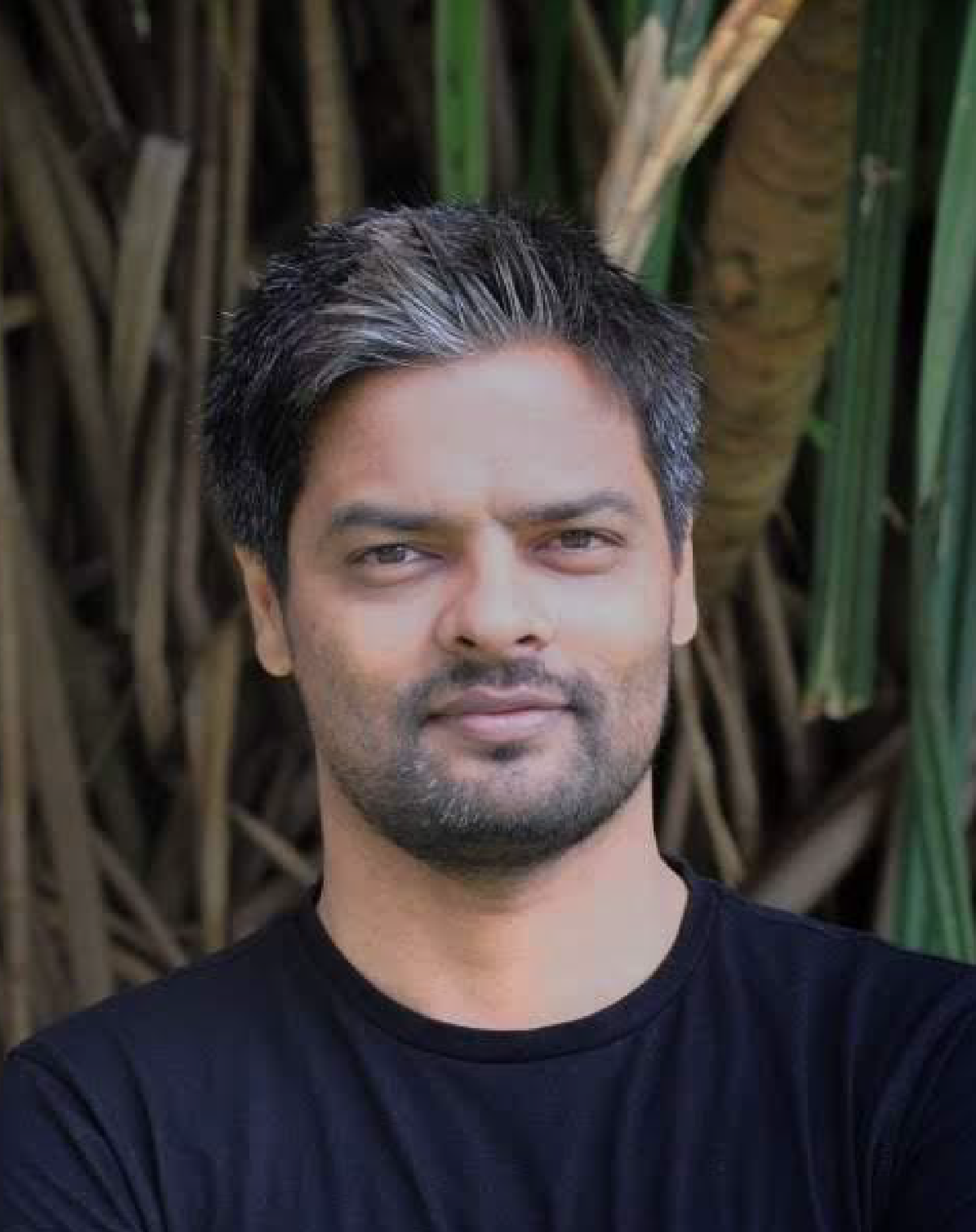 A man with short, dark, and slightly graying hair, wearing a black shirt, stands outdoors in front of tall grass or plants.