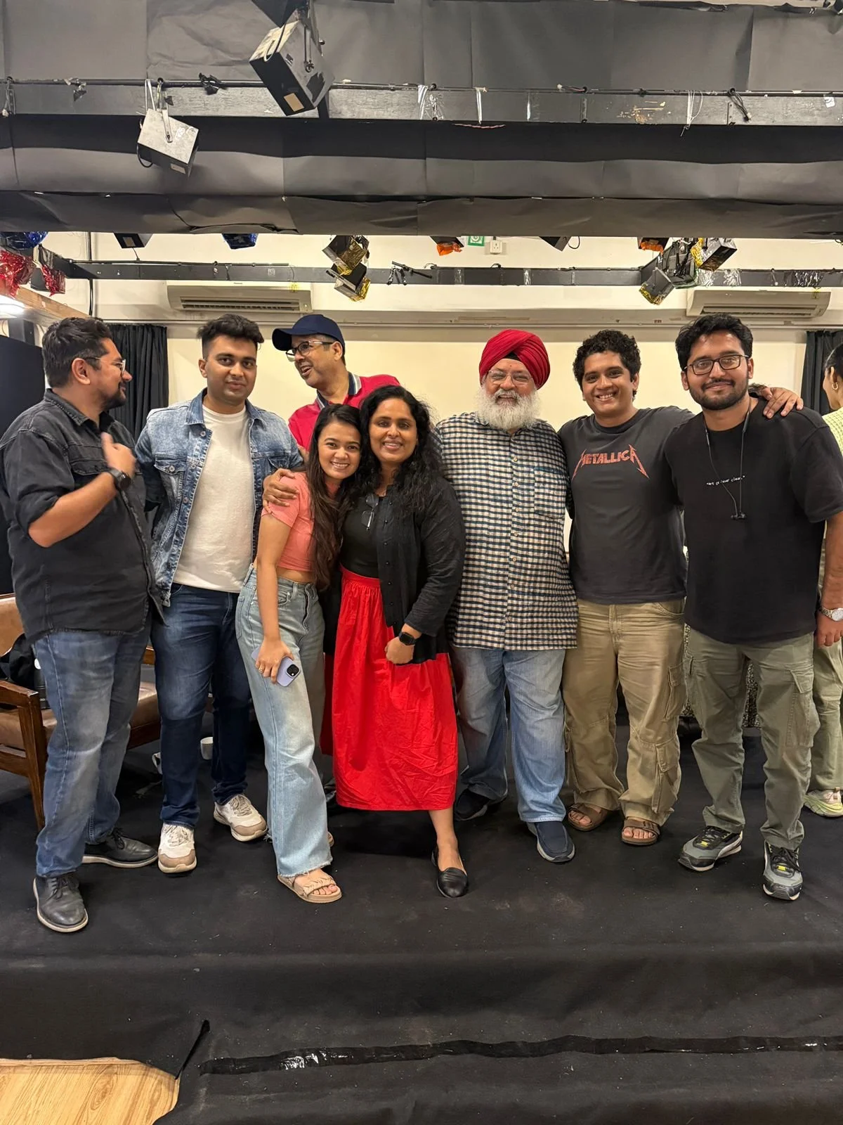 A group of eight diverse people standing together in a room, smiling and posing for a photo. There are five men and three women, with the man in the center wearing a red turban and a checkered shirt, and the woman in the middle wearing a red skirt and black top. They appear to be at an event or celebration.