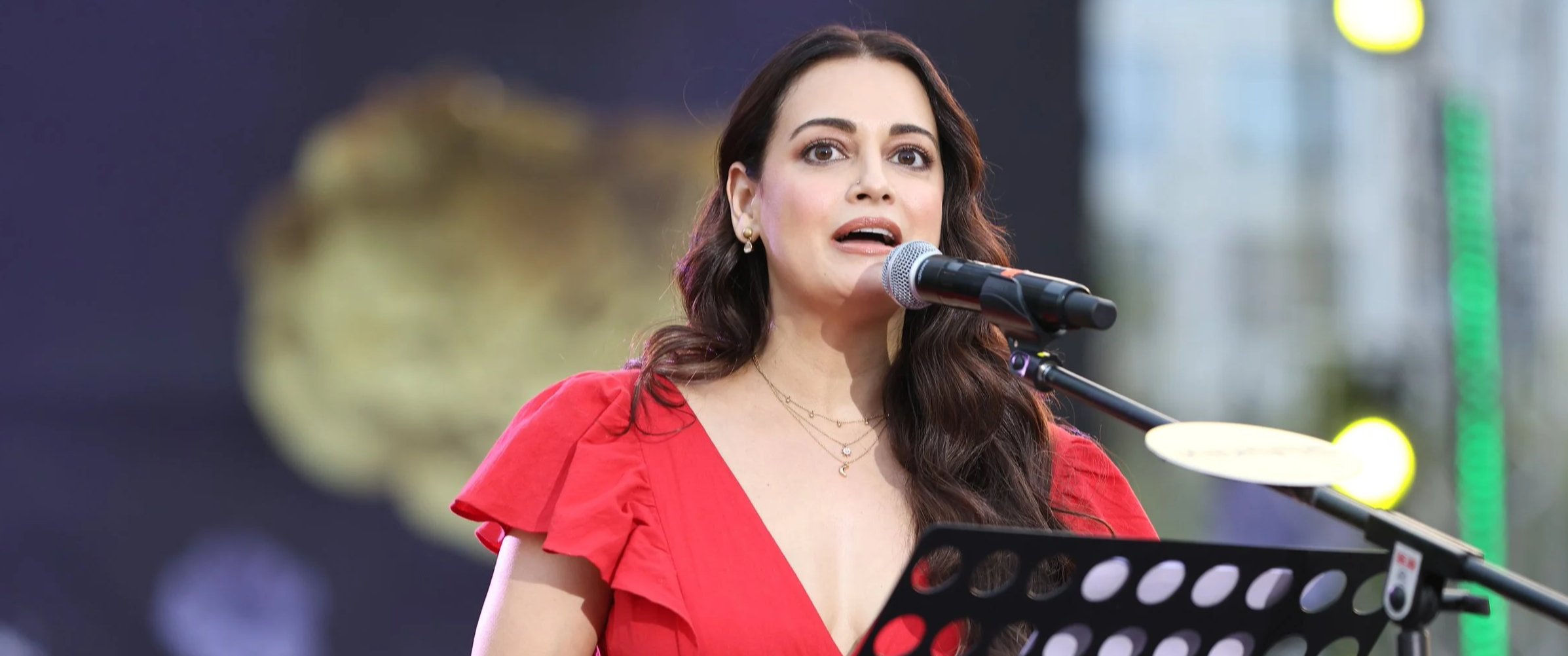 A woman (Dia Mirza) with long dark hair wearing a red dress speaking into a microphone on stage.