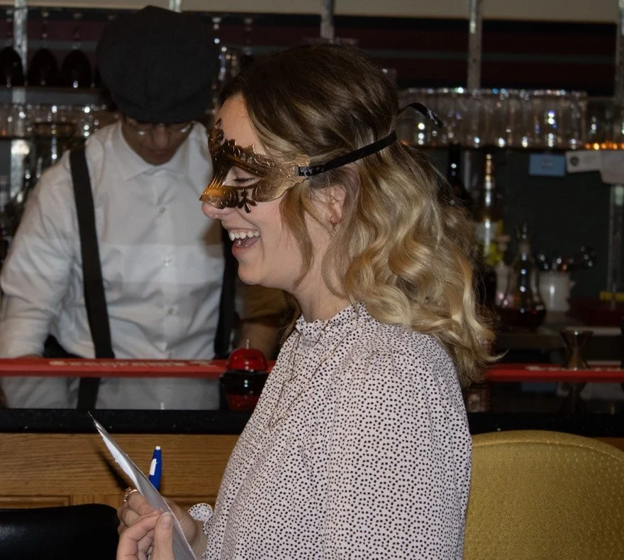 Woman wearing a masquerade mask and holding a card, smiling and laughing during a celebration, with a bartender preparing drinks in the background.