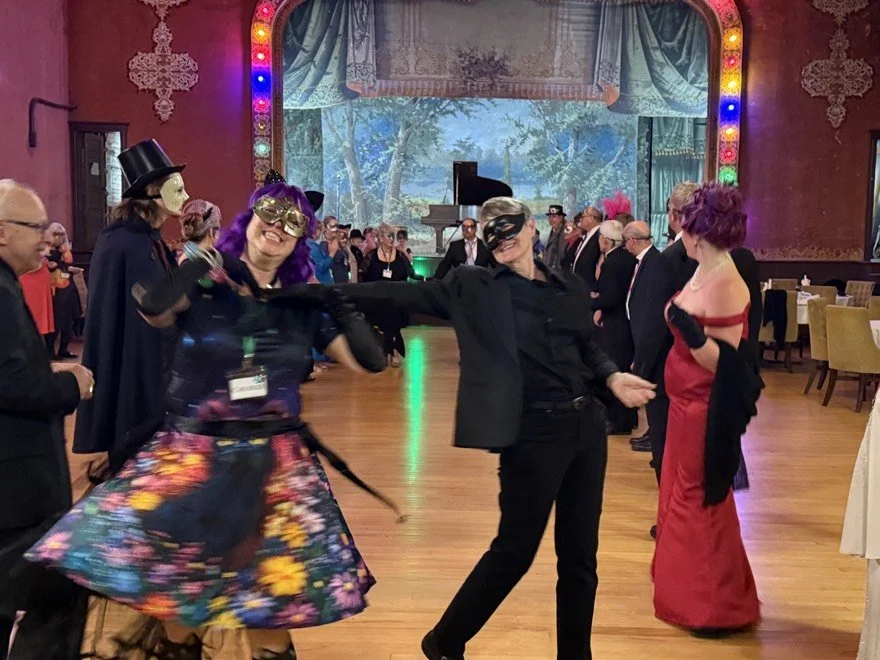 People at the masquerade dressed in costumes, dancing and socializing in a decorated venue with colorful lights, a stage with a scenic backdrop, and guests wearing masks and glamorous dresses. In the foreground, Chudlee is seen smiling and dancing. 