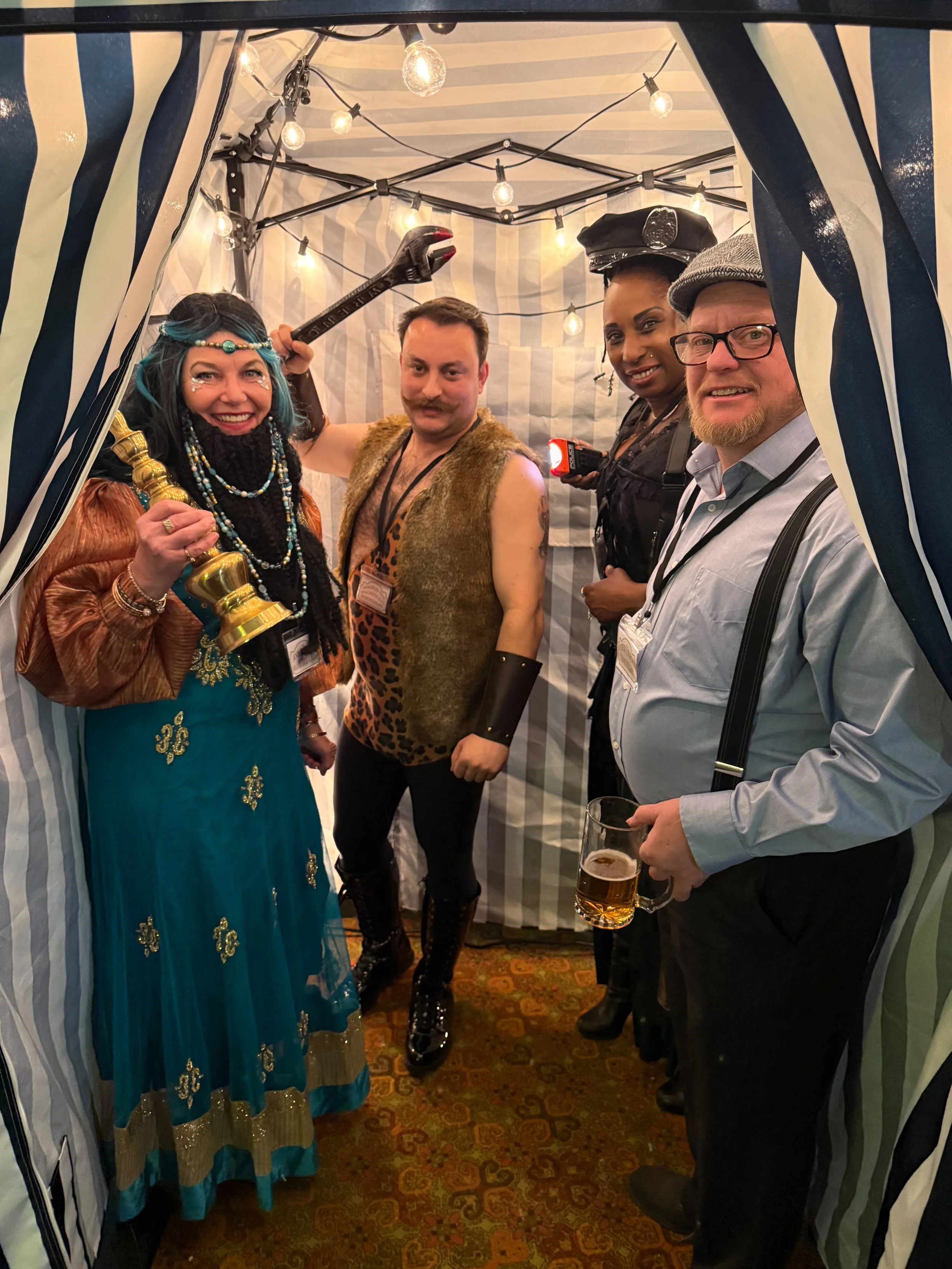 the bearded lady, strongman, ticket taker and unicyclist all together in a photo booth dressed in costumes, with the bearded lady holding a golden candlestick and a man holding a beer mug. The scene has string lights and striped fabric creating a fes
