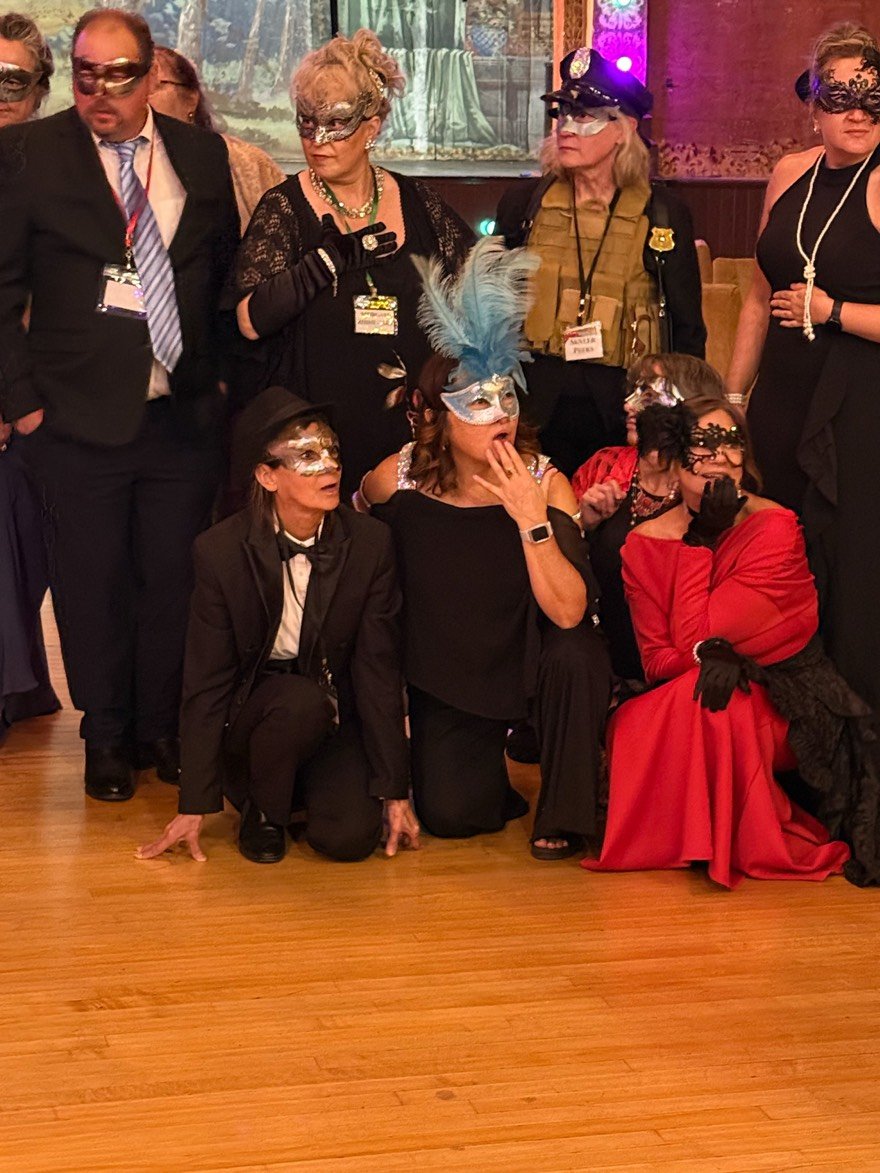 A gathering of guests look shocked while dressed in formal attire with masks, gathered in a ballroom with wooden floors and decorative backdrop, participating in a masquerade event.