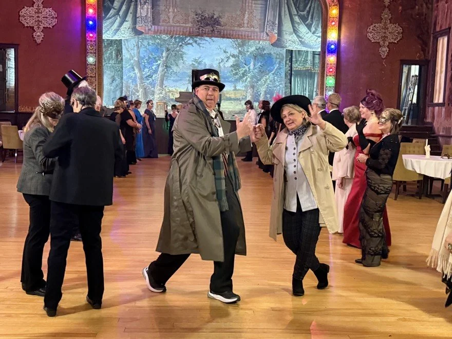 Detectives dancing and socializing in a decorated hall with colorful lights, a scenic backdrop, and tables at the Batcher Block Opera House.