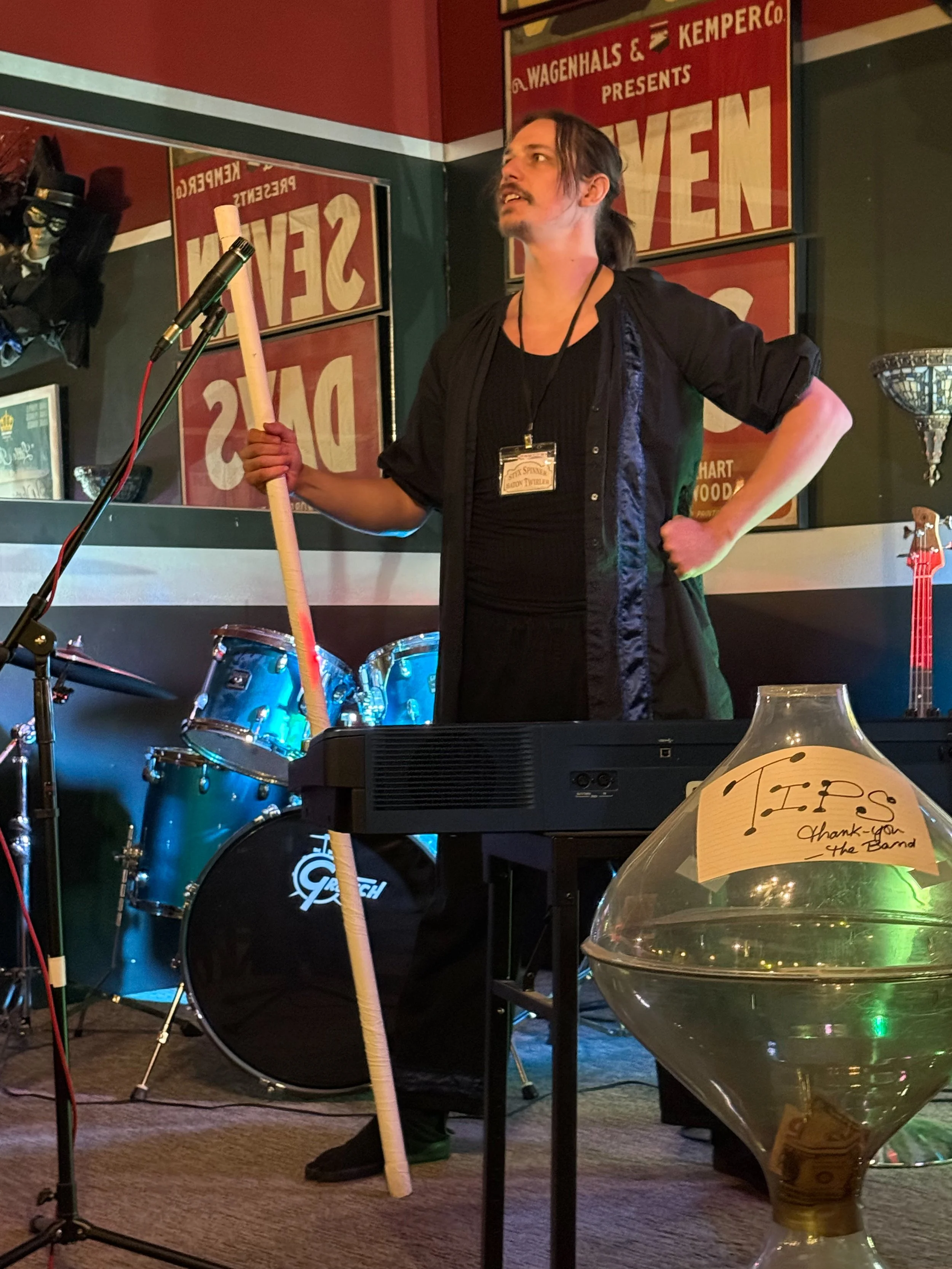 The Baton Twirler of the Velvet Noose Circus performs on a stage holding a wooden stick, wearing a black shirt and jacket, with drums and posters behind him.