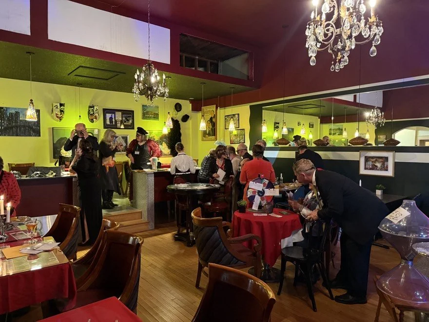 A busy hall filled with guests sleuthing and socializing in a colorful restaurant with yellow walls, chandeliers, and artwork.