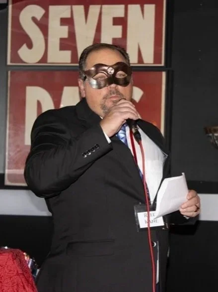 A man wearing a suit and a mask holding a microphone and reading from a paper on stage, serenading his love in the audience.