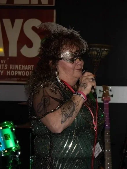T showgirl, with curly hair & a feathered mask, earrings, and a green dress, sings into a microphone on stage.