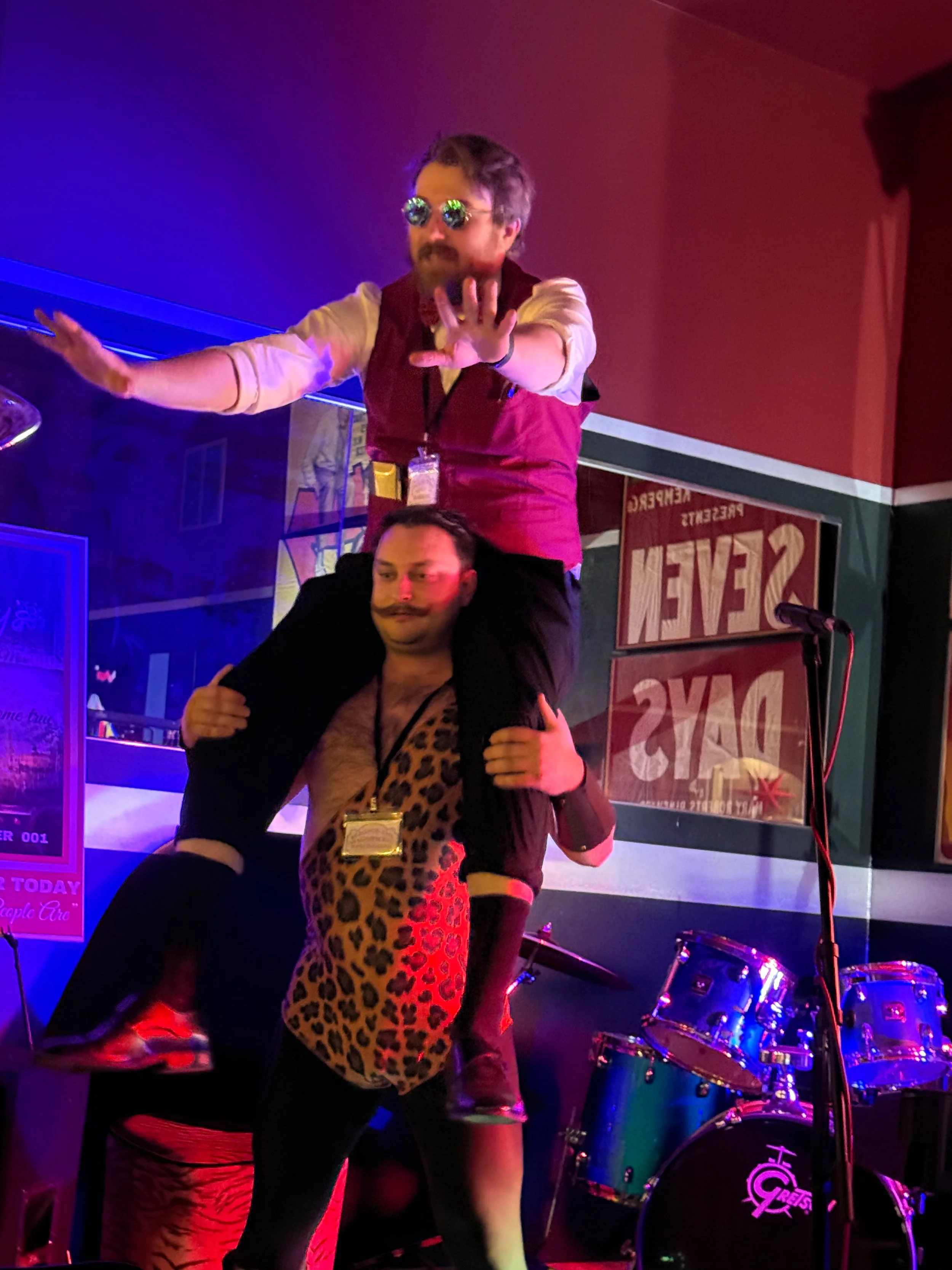 Two men, the strongman and the carnival barker of the velvet noose circus are performing on stage, one sitting on the other's shoulders, in a bar or club with drums in the background and colorful lighting.