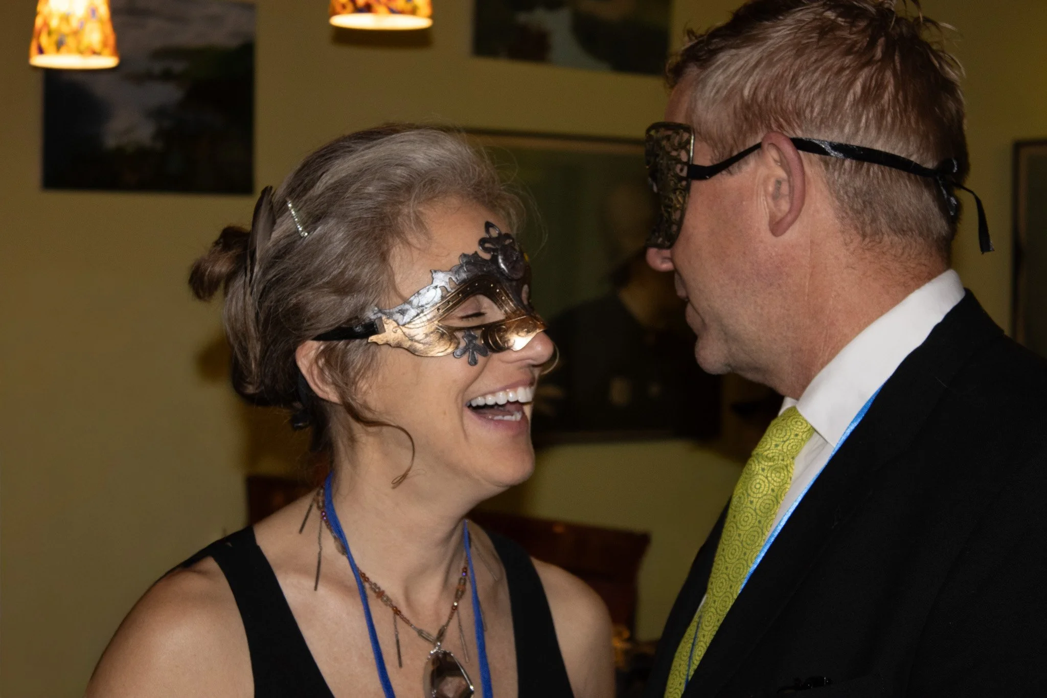 A woman and a man wearing masks are facing each other at a masquerade party, smiling and laughing over a shared secret.