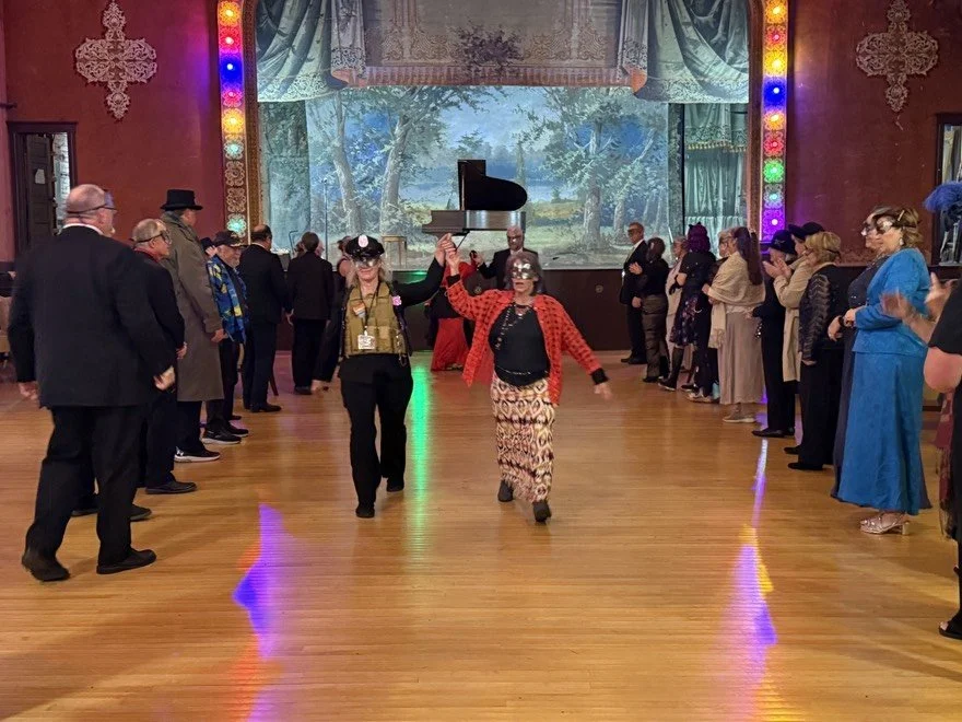 People dancing in a decorated ballroom with colorful lights, a wooden floor, and a backdrop painting of trees and sky. One is dressed as an officer in a bullet proof vest and hat. The other in elegant attire.
