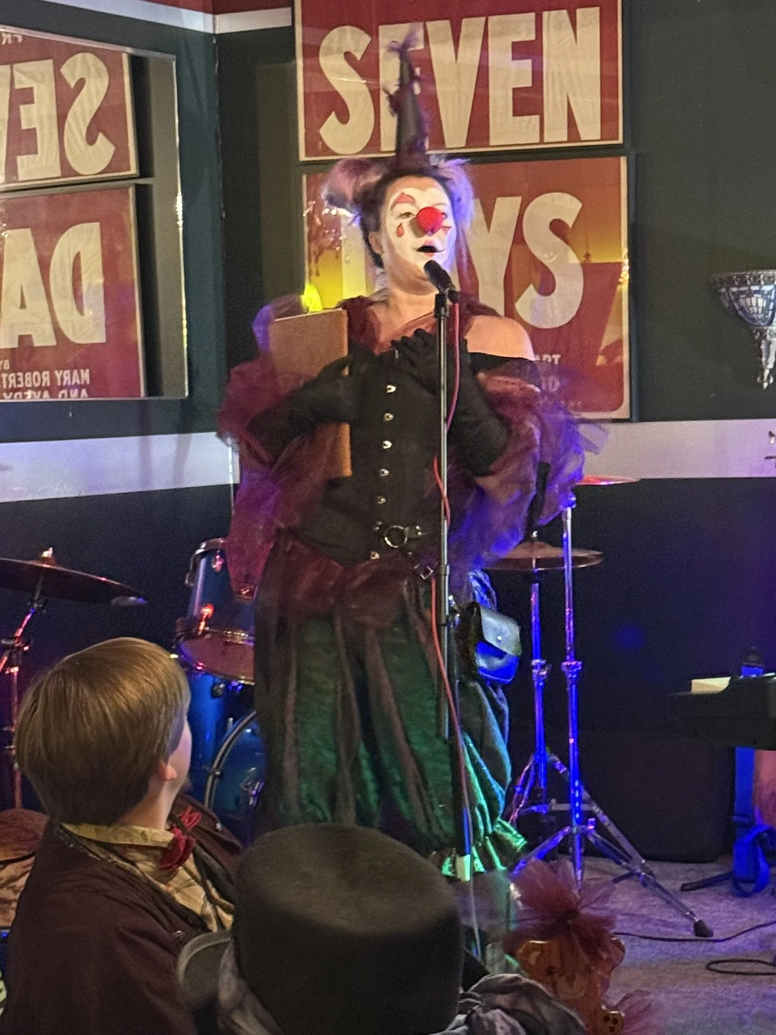 The Murder Mystryss dressed as a burlesque clown standing on stage in front of a microphone, mid-speech. She has white face clown makeup and a red nose with a pointy hat with pompoms. The outfit is burgundy, black, and purple.