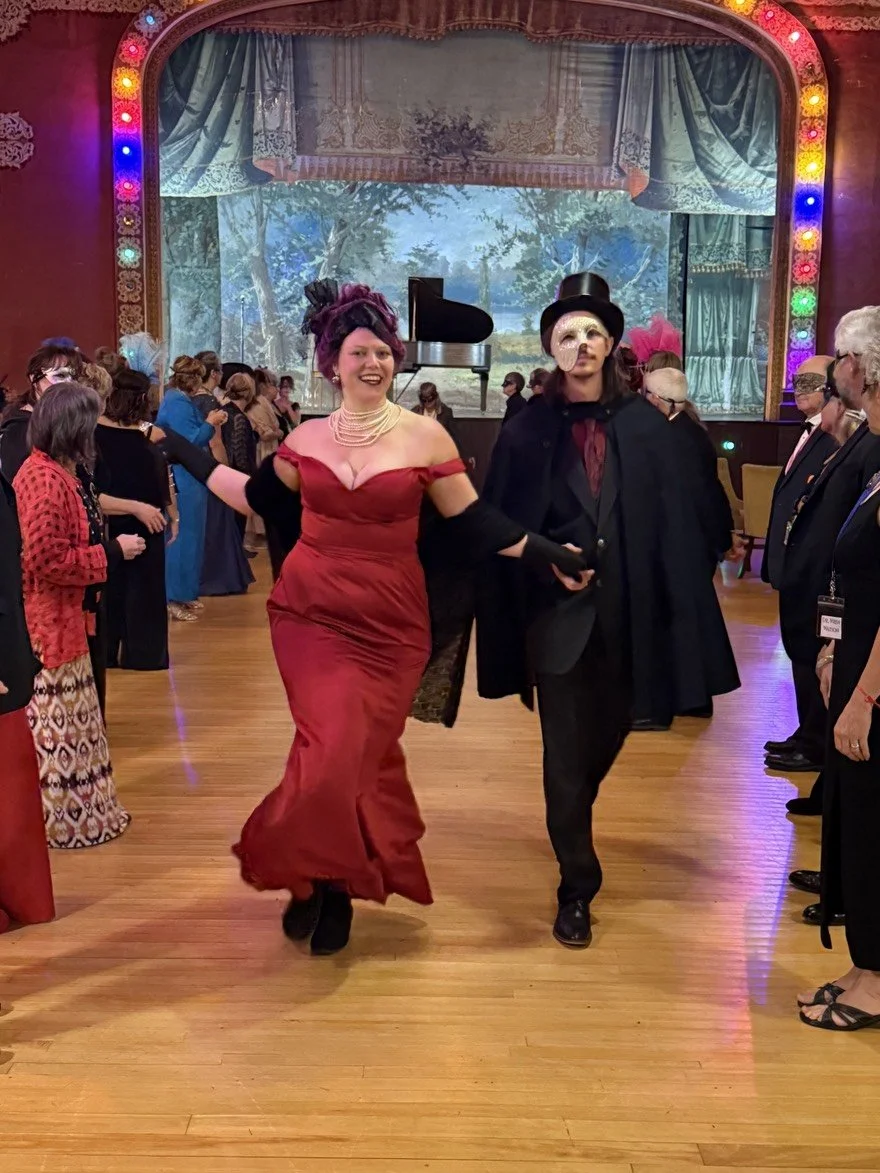 The Murder Mystryss, in a red gown and the Phantom, in a blue cape, dance through 2 lines of guests in a theater setting with a painted backdrop of trees and a piano, surrounded by an audience, some wearing masks.