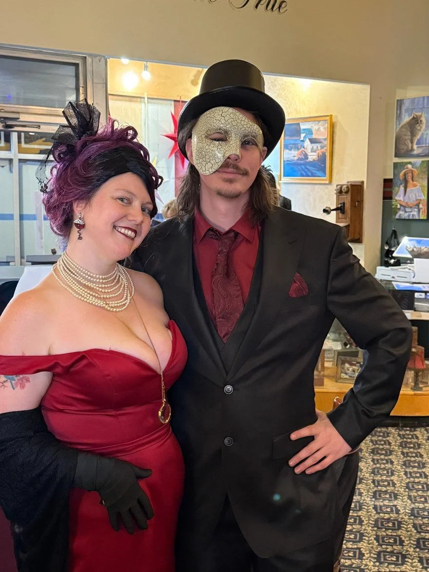 The Murder Mystryss in her red dress and pearl necklace, with purple hair and black gloves, smiling and posing with the Phantom of the Batcher Block Opera House in a black suit, red shirt, and black top hat, wearing a cracked mask over one eye, in Fr