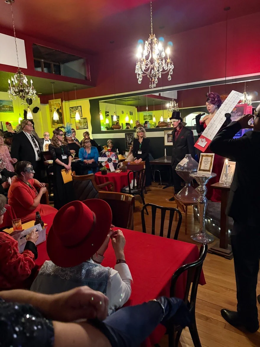 People attending a themed event or performance at a restaurant with red and green walls, chandeliers, and framed pictures. Some attendees wear costumes, including a woman in a red hat and sequined jacket. Others are watching the Mystryss on stage.