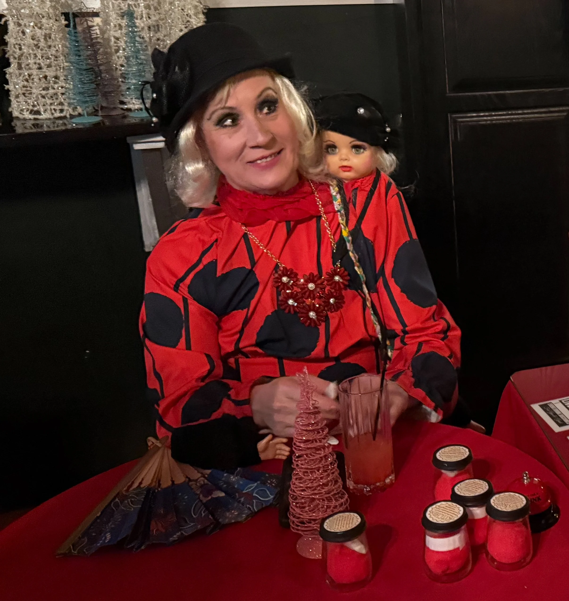 Someone in a two-headed woman costume sits at a table with various jars on it. The woman has blonde hair and makeup, and the setting appears festive with holiday decorations on the table and in the background.