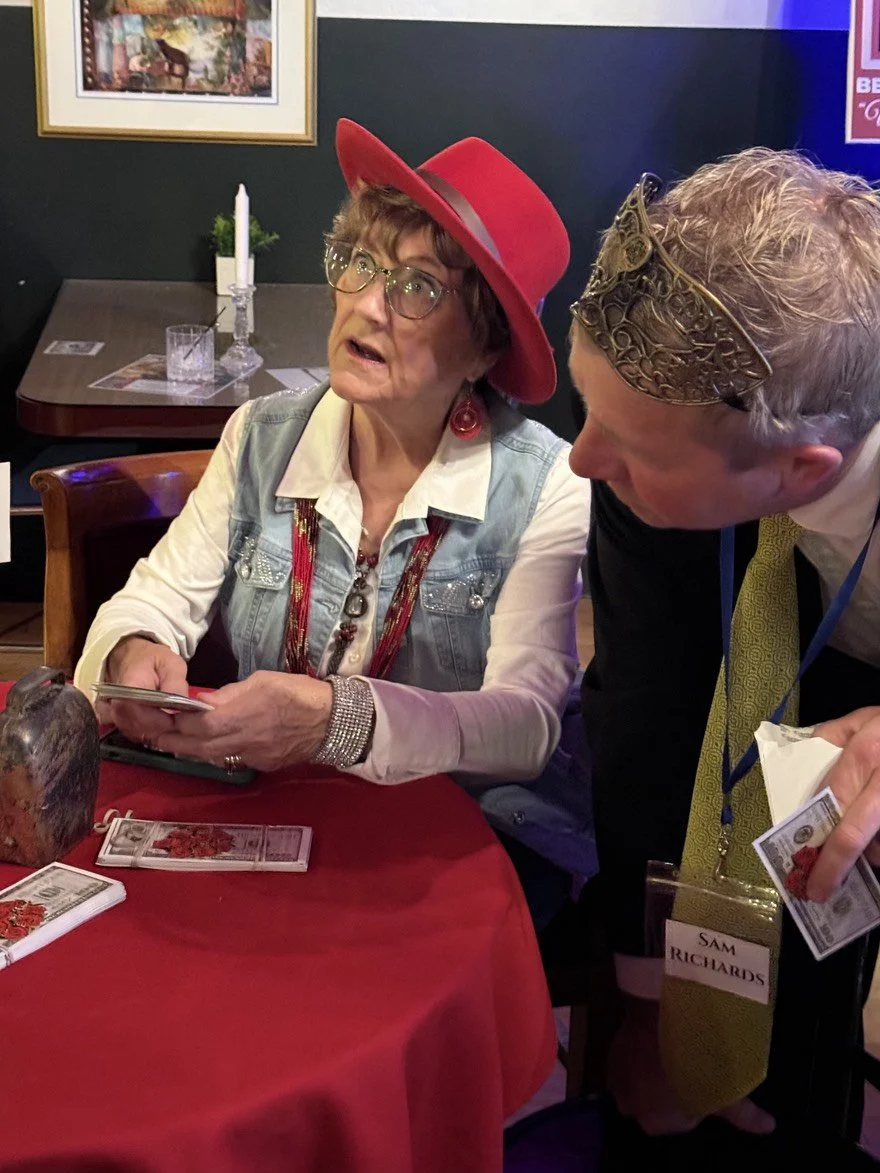 A woman wearing glasses, a red hat, and a denim vest sitting at a table counting money. A man wearing a gold mask, a yellow tie, wearing a name badge, leaning toward her. The table has a red tablecloth, and there is a candle, a small potted plant, an