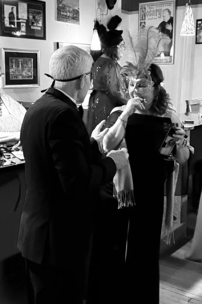 A black-and-white photo of a masked man in a tuxedo speaking with a laughing woman wearing a decorative mask and a feathered headdress at the Murder at the Masquerade Mystery Event.