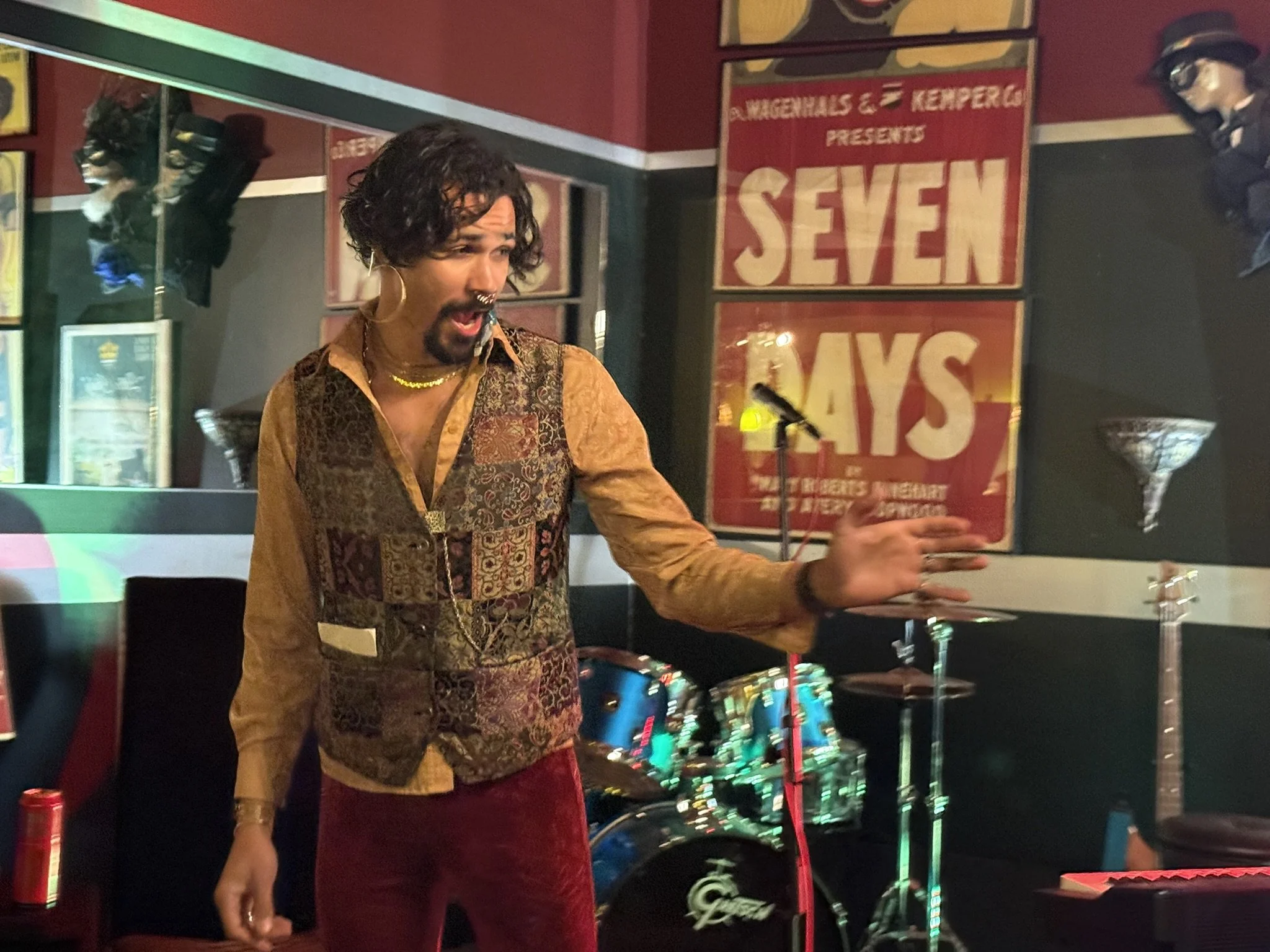 The escape artist, with curly dark hair performs on stage, arms outstretched, wearing a multicolored patterned vest, gold shirt, red pants, and hoop earrings, with a septum piercing. The background features a drum set, posters, and decorative lightin