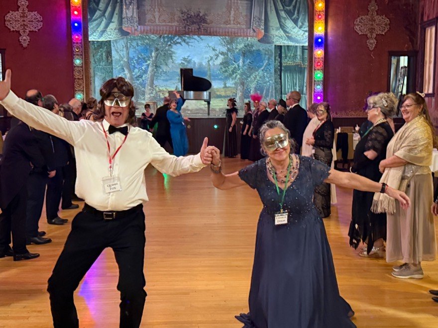 People wearing masquerade masks and formal attire, dancing and having fun at a lively indoor party with a decorated ballroom and a grand piano in the background. A couple dances center stage. Her in a blue gown, him in a waiters uniform and wig.