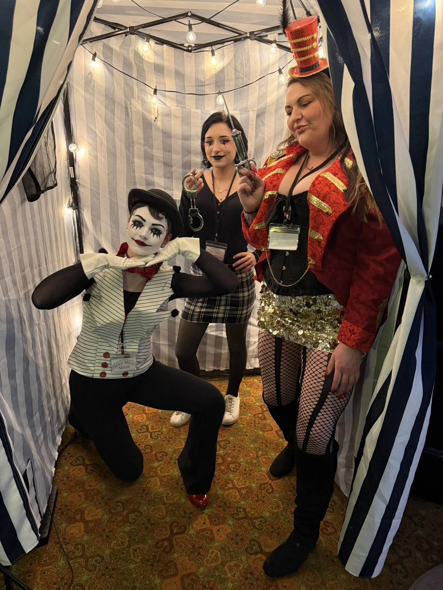 The mime, ringmaster's assistant, and contortionist in circus or carnival costumes inside a striped tent with string lights. The person on the left is dressed as a mime with white face paint, black makeup, and a vintage outfit. The contortionist in t