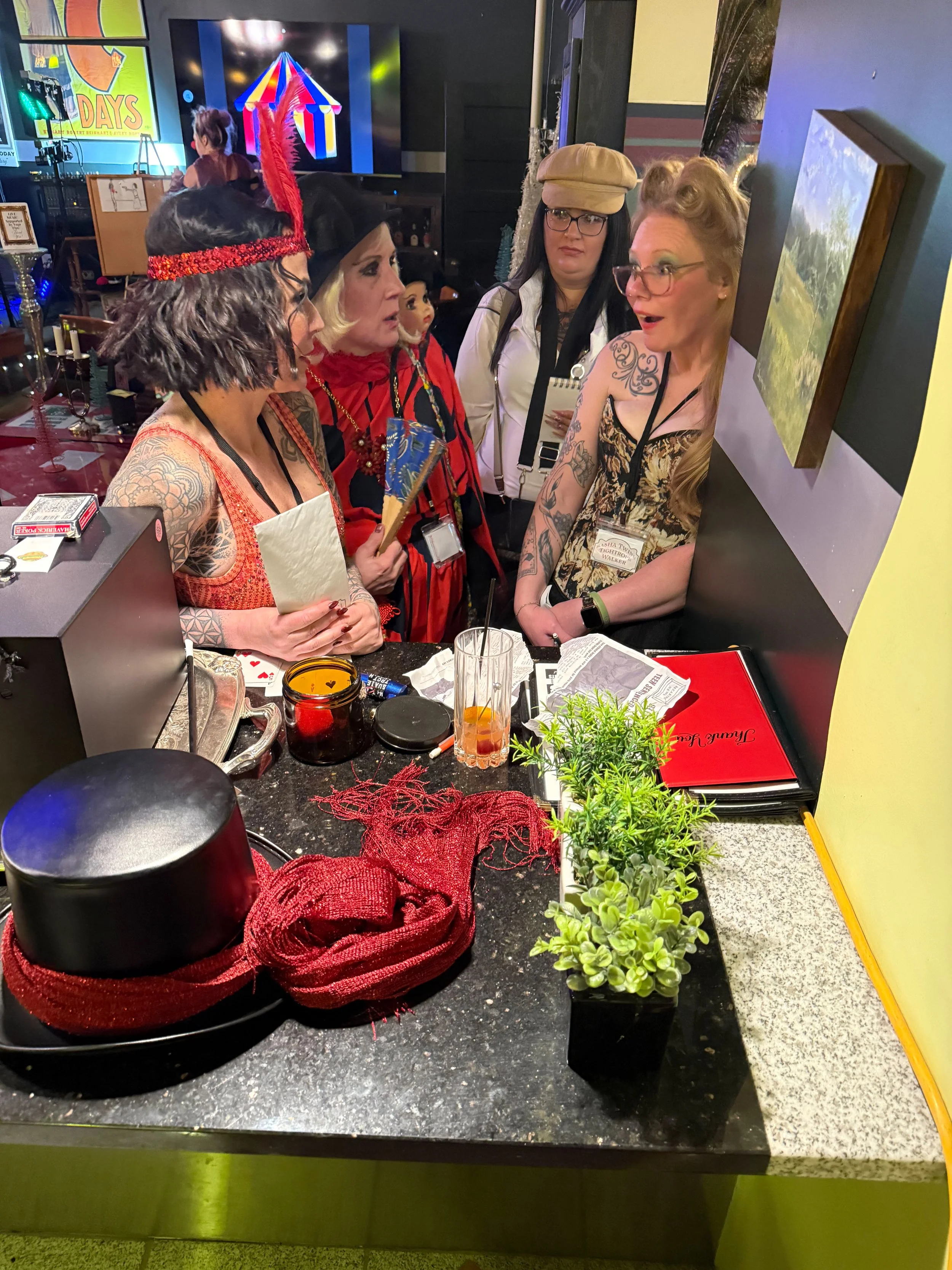 The trapeze artist, two-headed wonder, detective and tightrope walker, all dressed in costumes and accessories are engaged in a conversation at a bar or reception area, with party decorations, a plant, and various puzzle and clue related objects on t