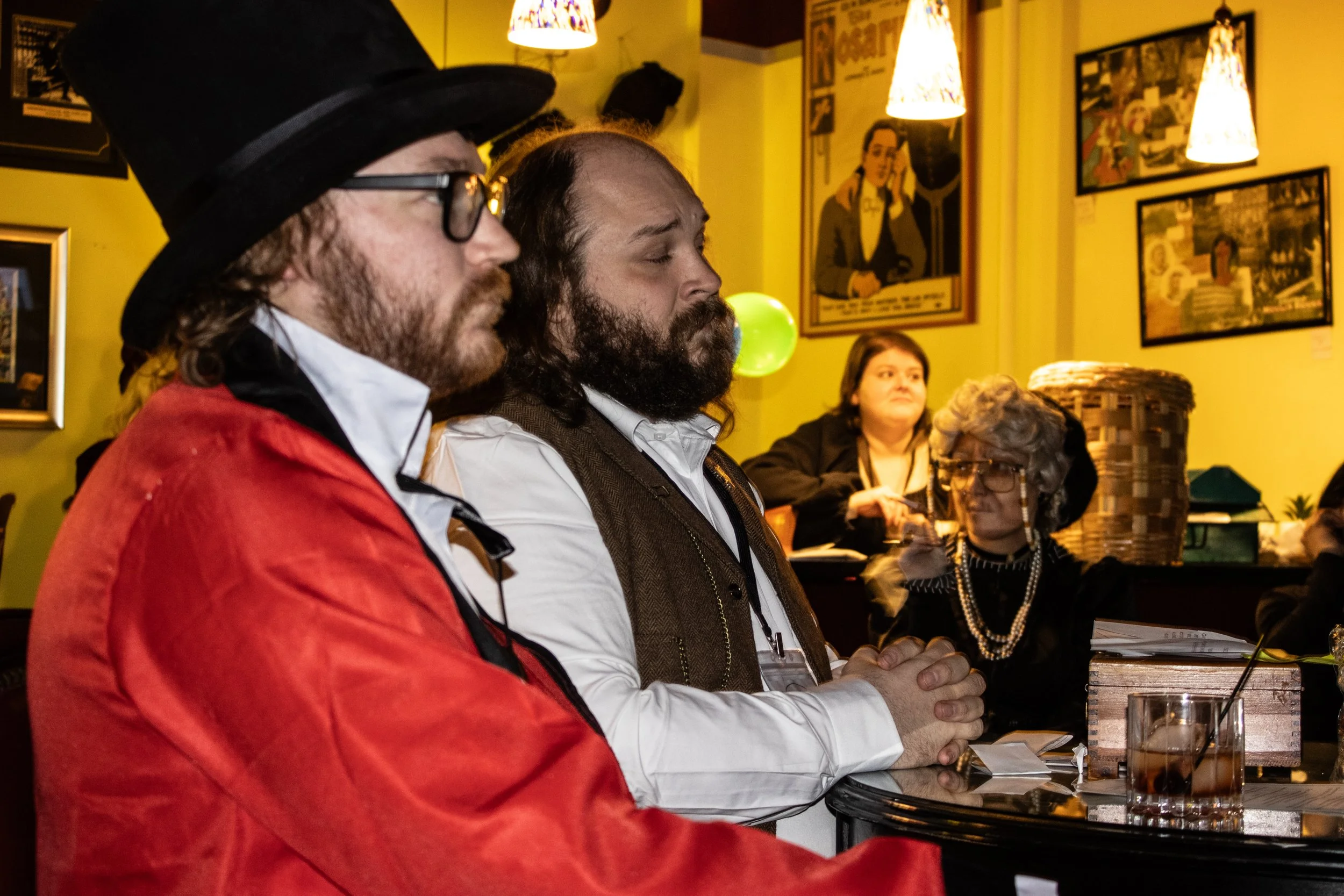 The ringmaster and talent scout are sitting at a table in a cafe, listening attentively. The ringmaster in the foreground wears a red jacket and a black hat, the talent scout next to him has long hair and a beard, and the caricature artist sits behin